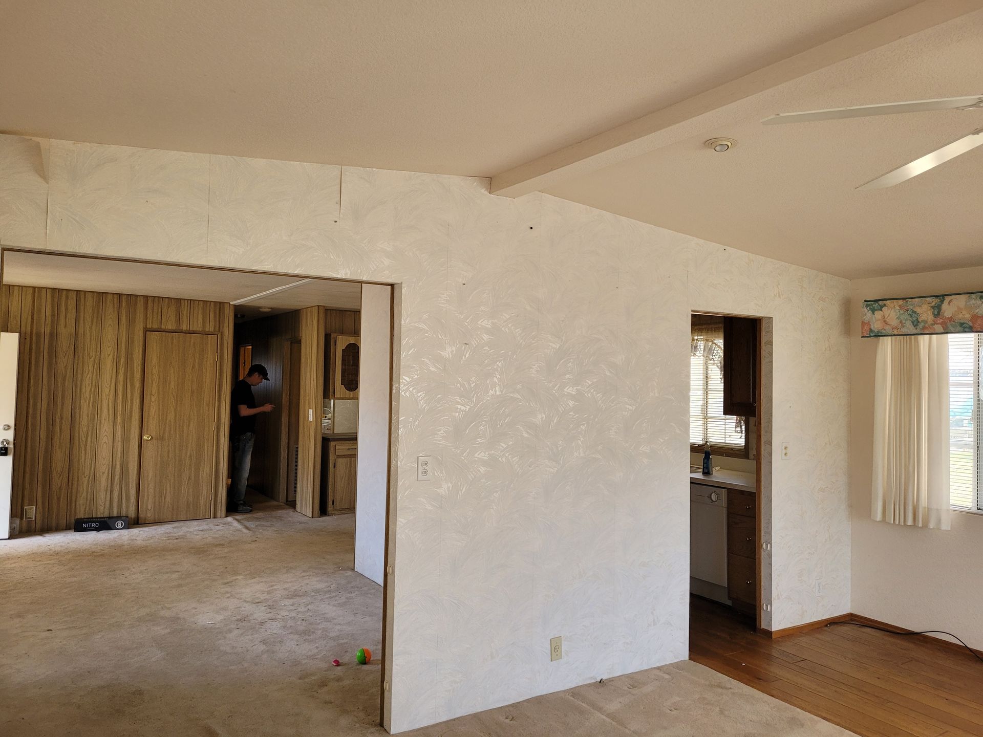 Interior view of a room under renovation. A person stands in a doorway, with exposed wall studs and peeling wallpaper visible.
