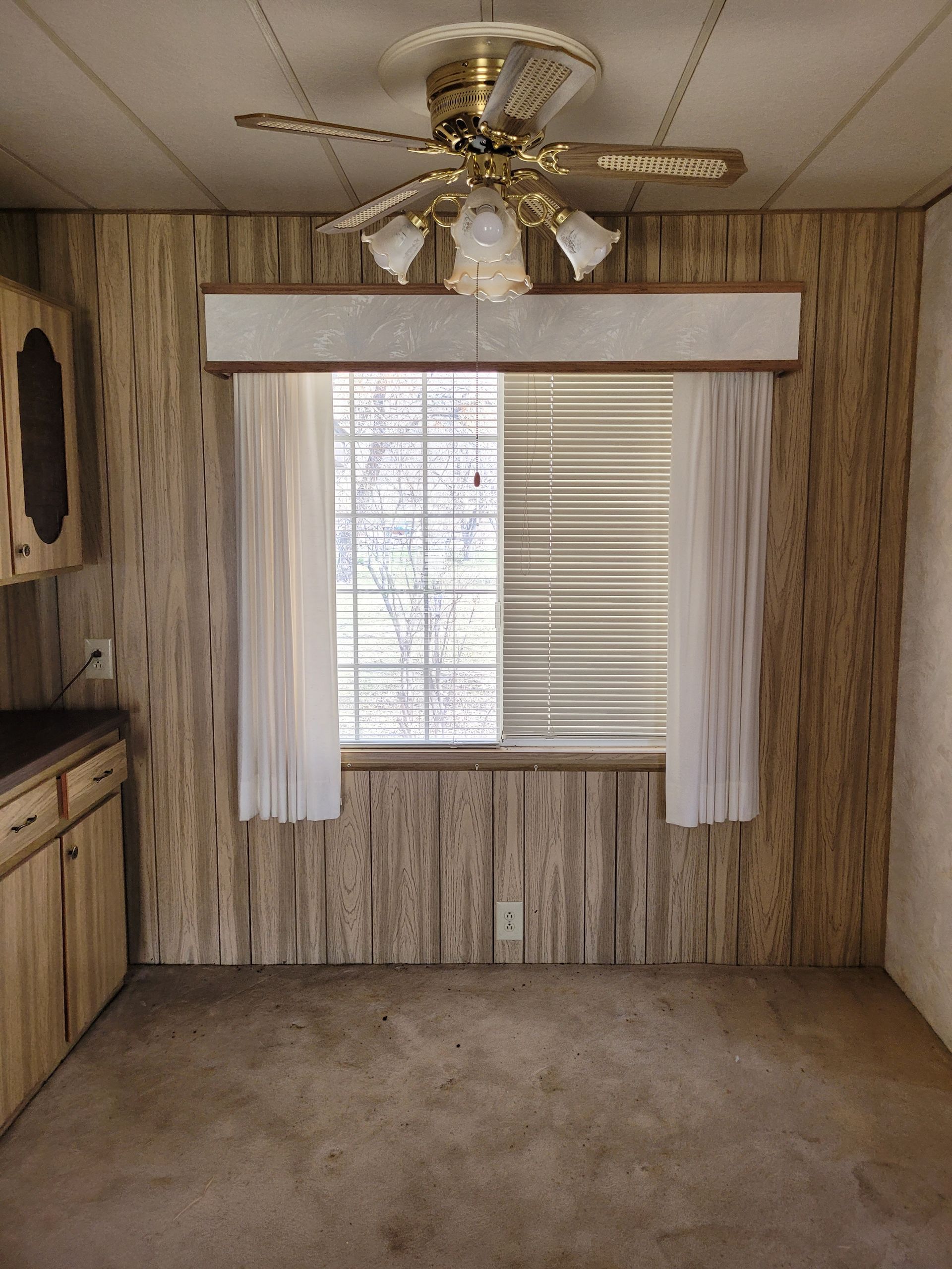 Interior shot of a room with wood paneling, a window with sheer curtains and blinds, and a ceiling fan. The carpet is worn.