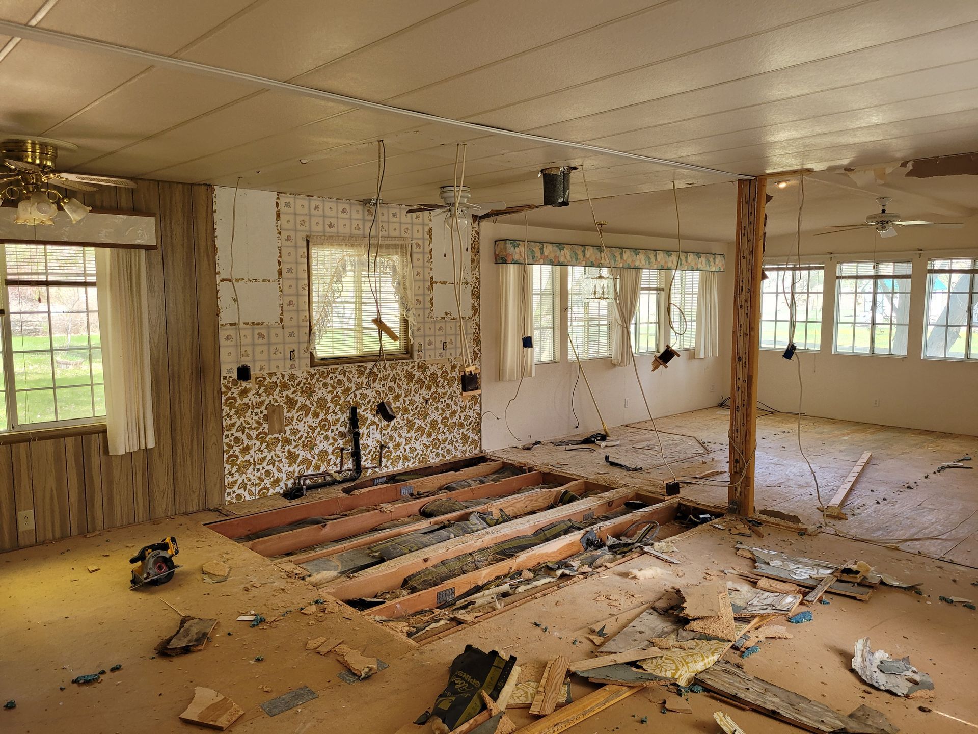 Interior of a dilapidated room under renovation. Exposed floorboards, damaged walls, debris, and two ceiling fans are visible.