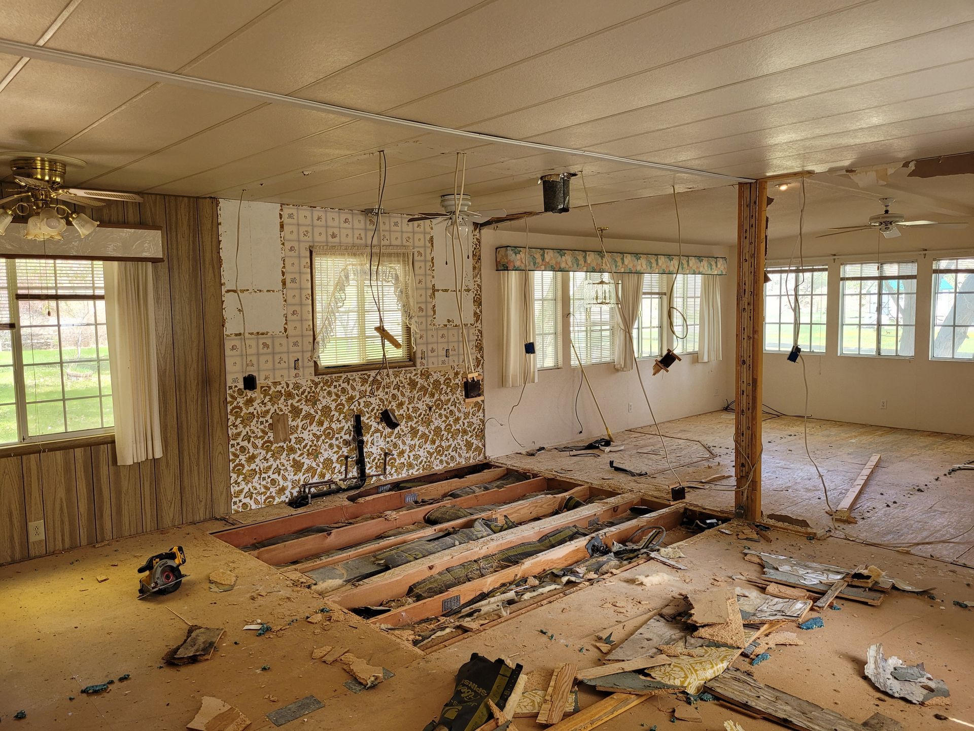 Interior room undergoing demolition. Damaged flooring, exposed framing, and debris are scattered throughout. Windows and a fireplace are visible.