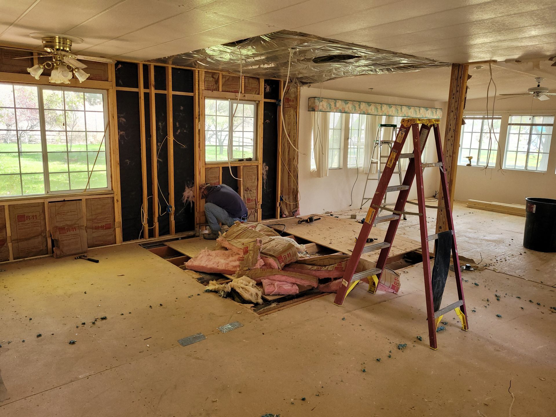 Interior of a room undergoing renovation. A person works on a wall with exposed studs and insulation. Debris and a ladder are in the space.