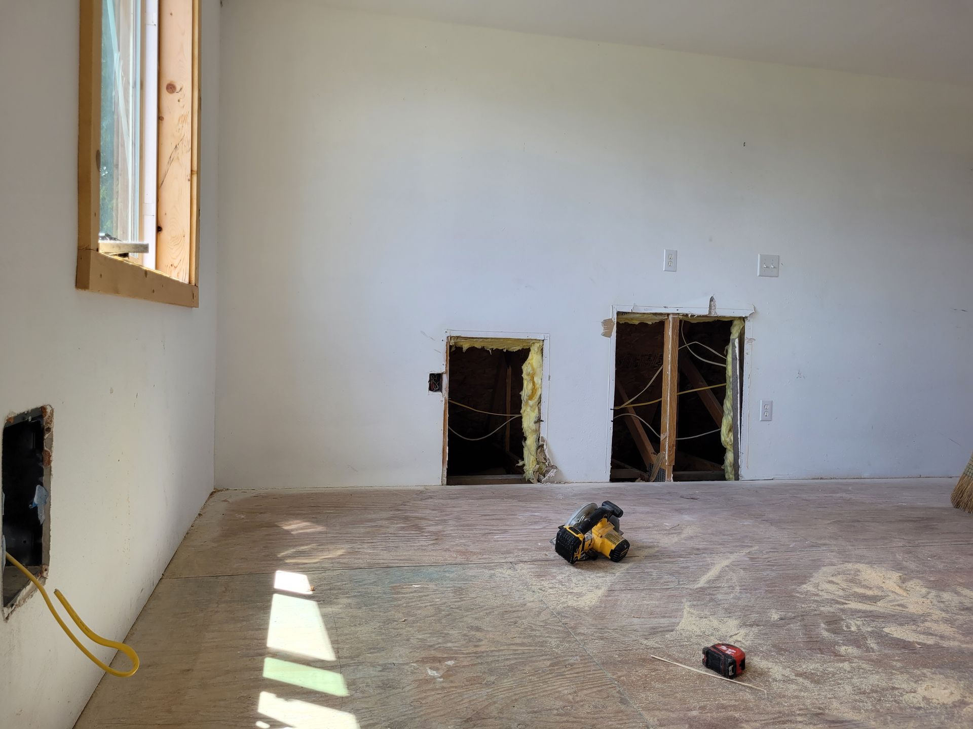 Interior view of a room under construction. Two rectangular holes in the wall, exposed wiring and a window with a wooden frame.