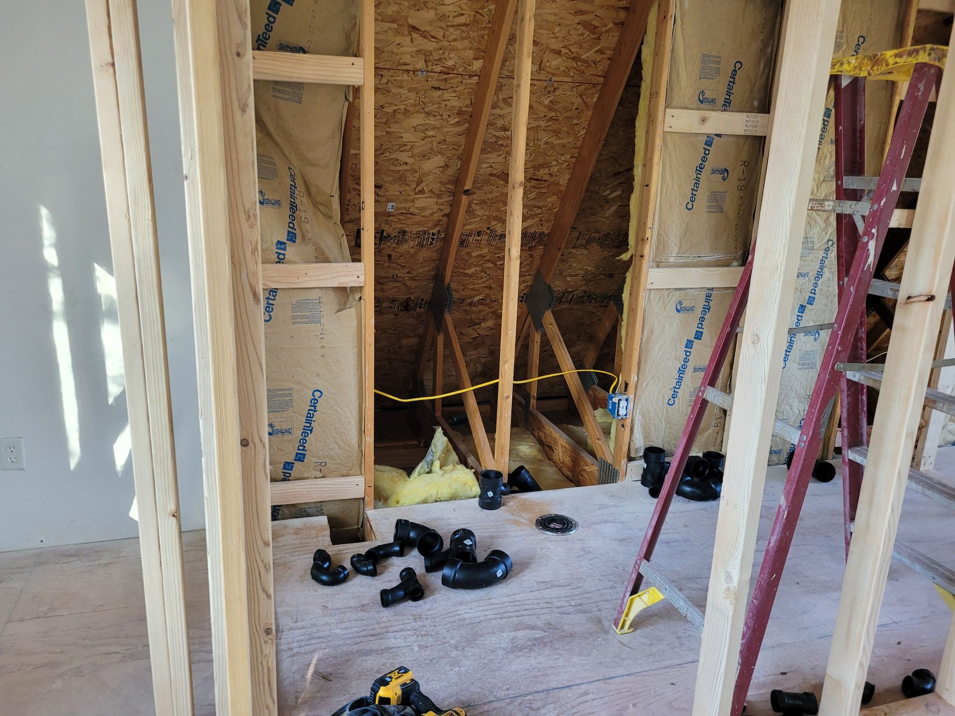 Interior view of a room under construction, with exposed wooden framing, insulation, and plumbing, next to a ladder.