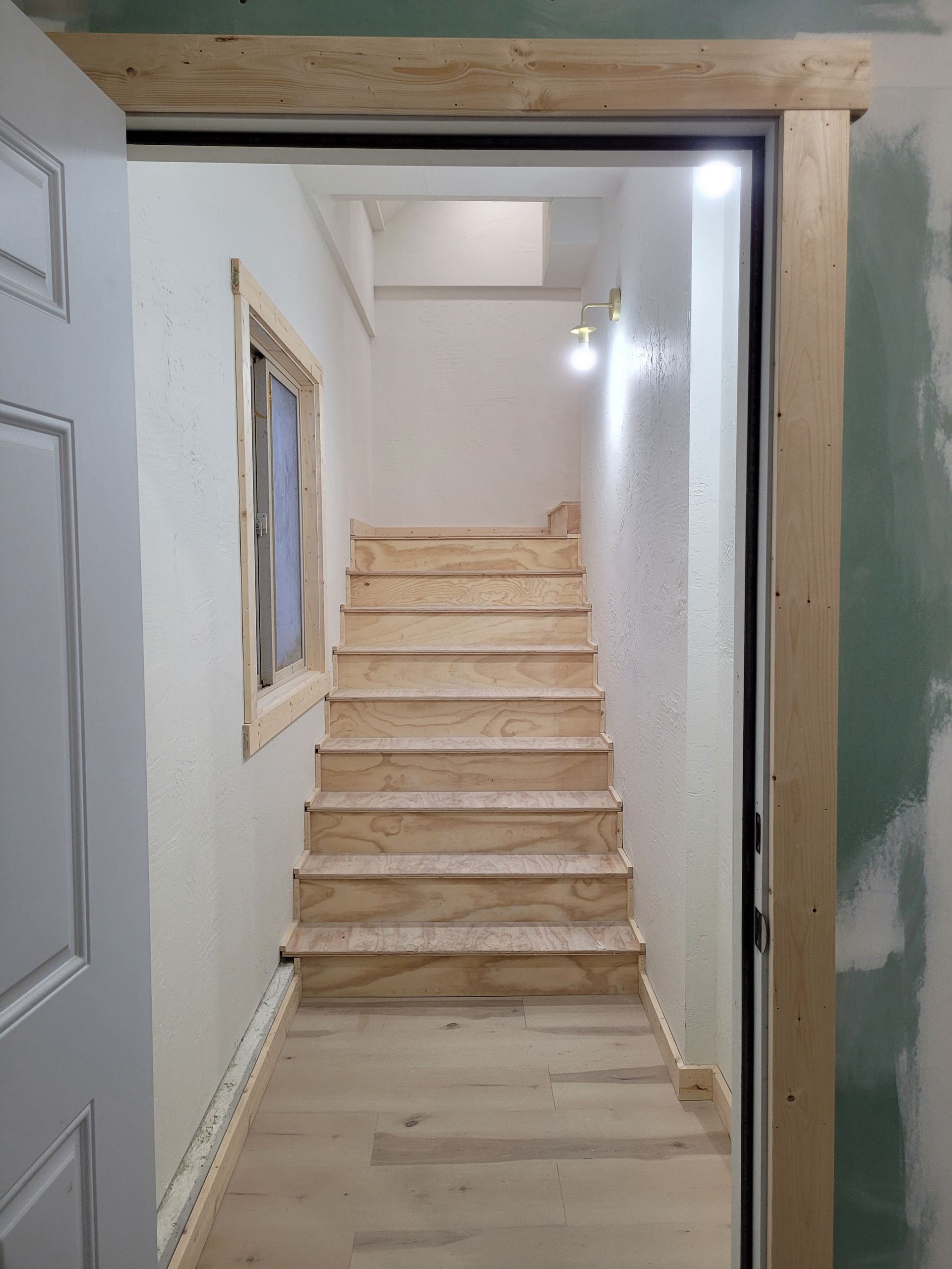 Wooden staircase leading upwards, viewed from a doorway. Light-colored wood steps, walls, and flooring.