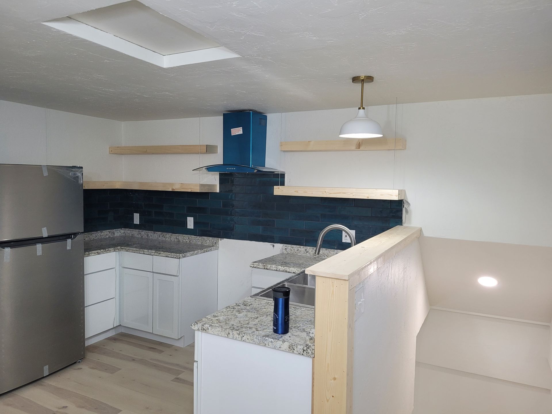 A small, newly renovated kitchen with white cabinets, light wood floating shelves, and a blue tile backsplash.