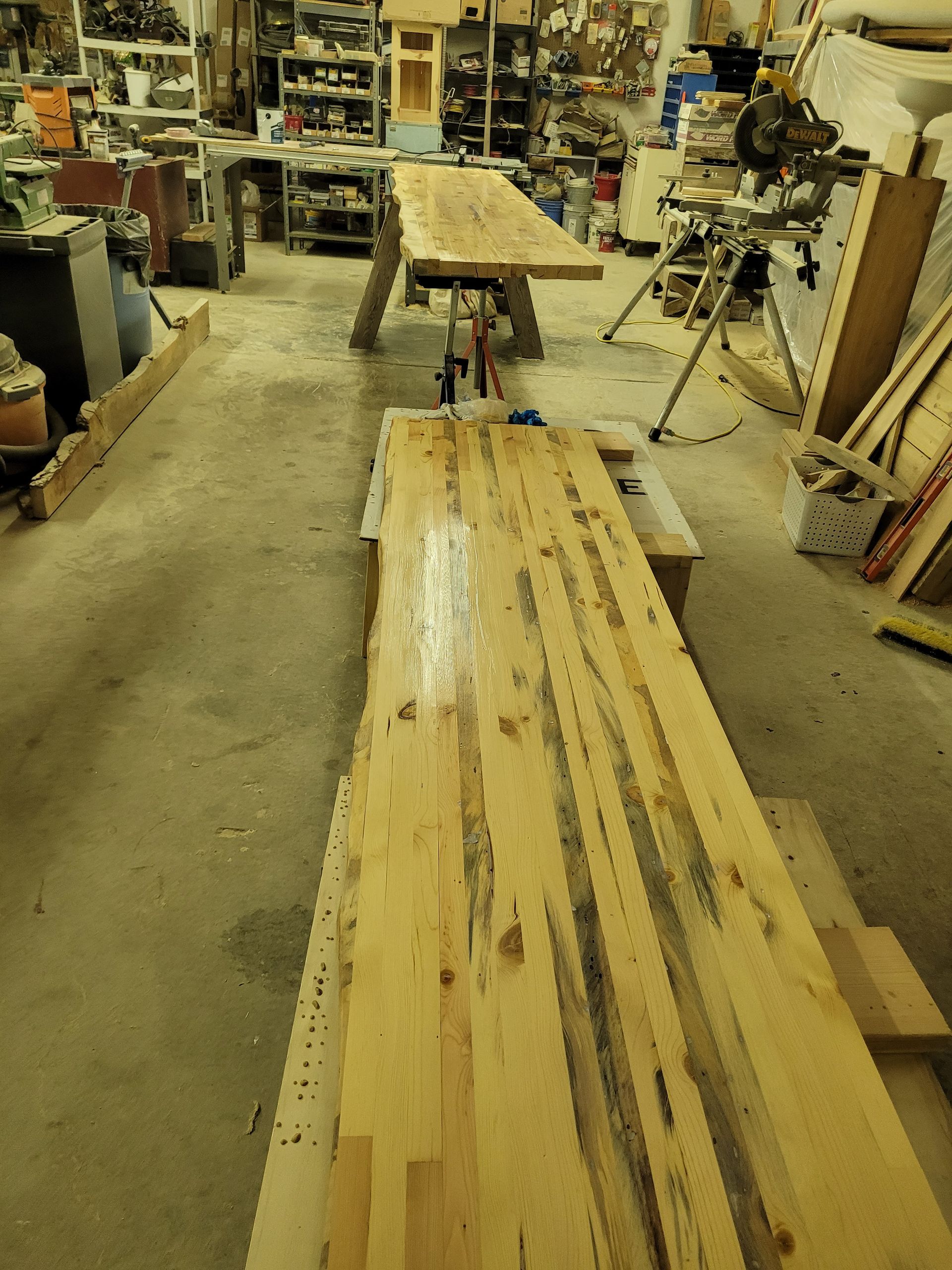 A workshop with two large wooden tables, one in use, and a long unfinished wood surface on the floor. Tools and equipment are visible.