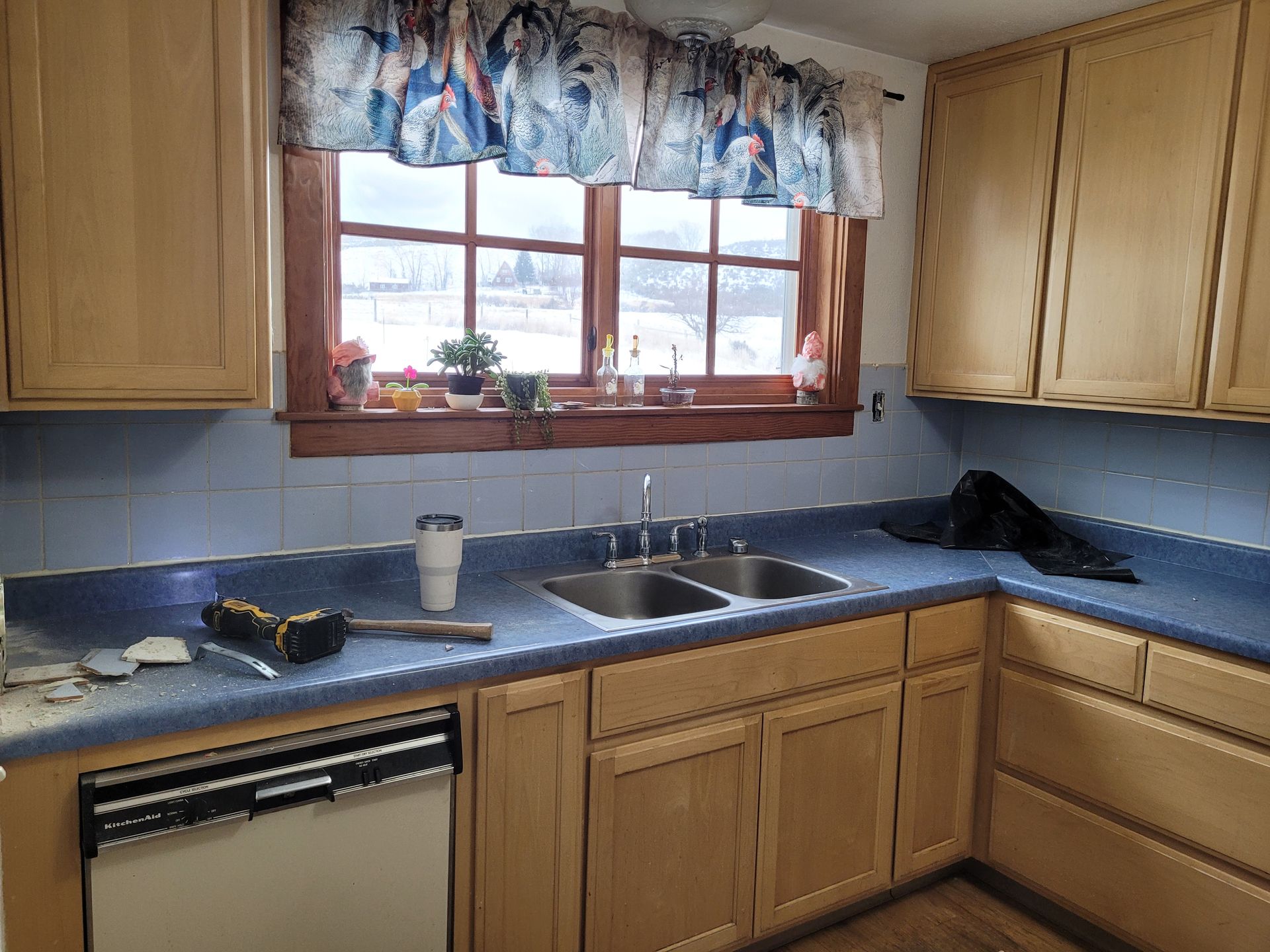 A kitchen with light wood cabinets, a blue countertop, and a window above the sink. A dishwasher and some tools are visible.