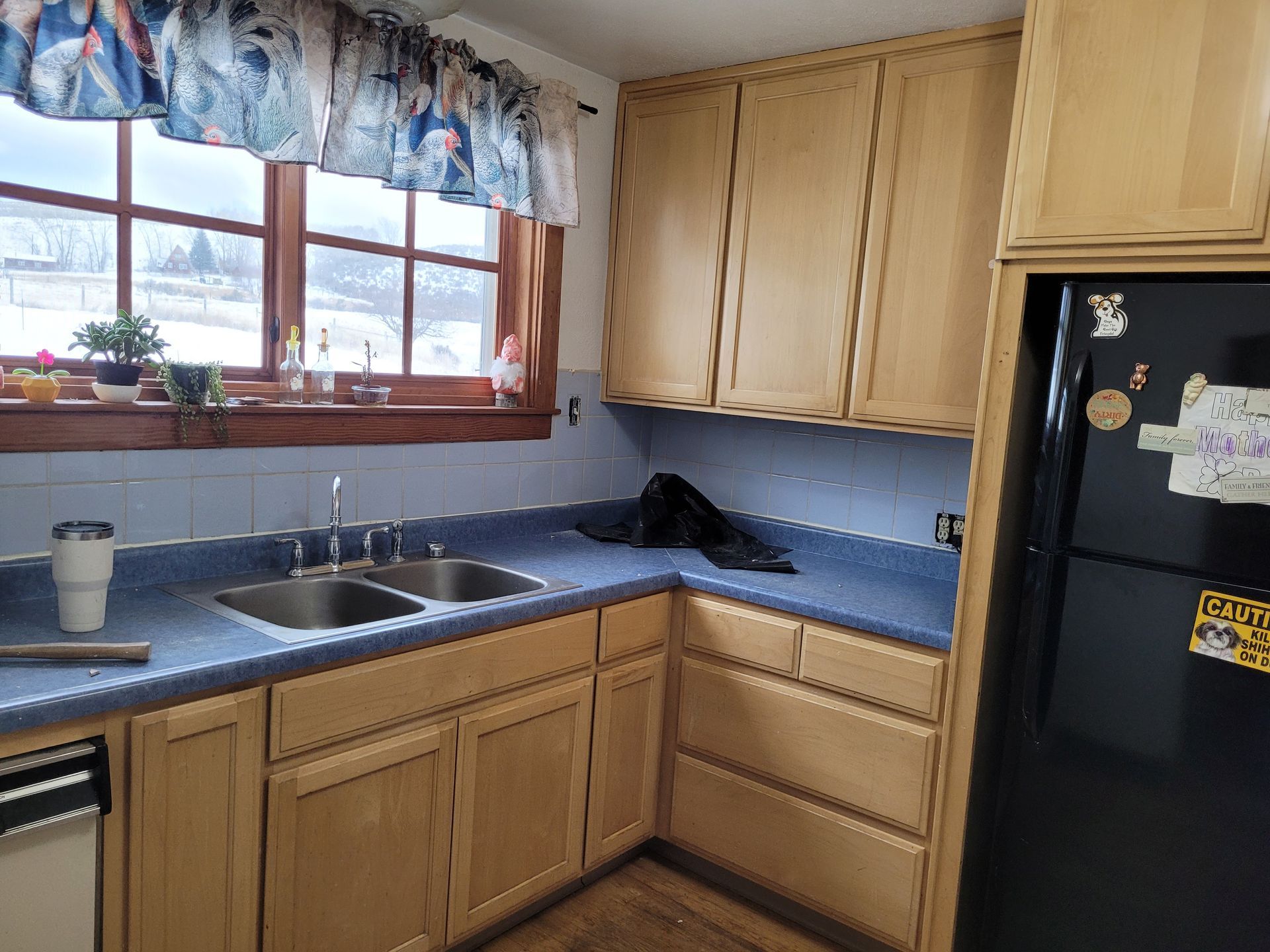 A kitchen with light wood cabinets, blue countertops, and a window above the sink. A black refrigerator is on the right.
