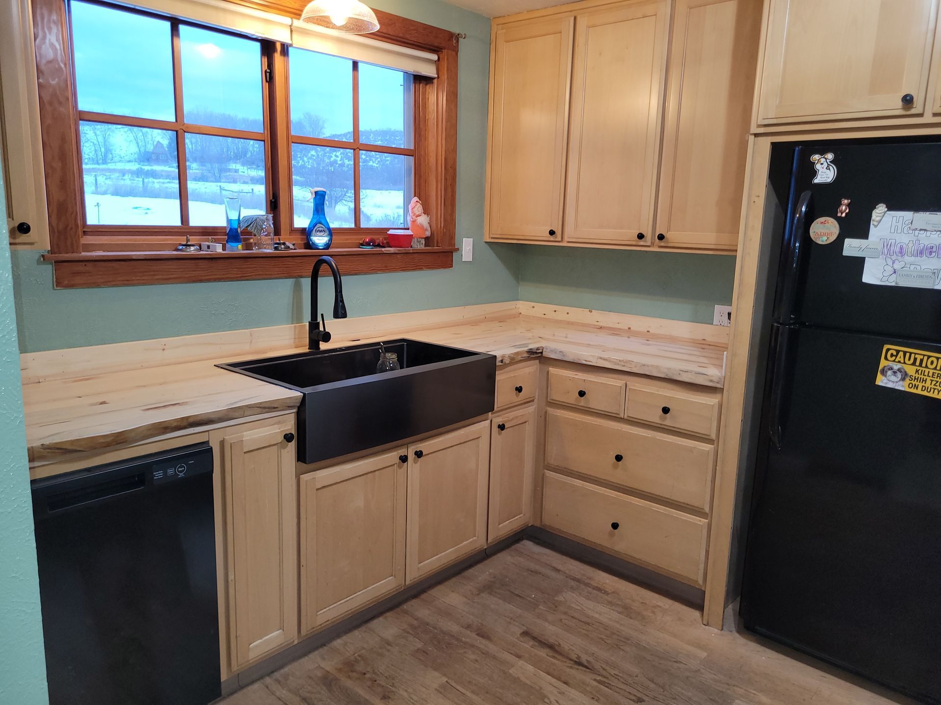 A kitchen with light wood cabinets, a black farmhouse sink, and a black refrigerator. The walls are painted a pale green.