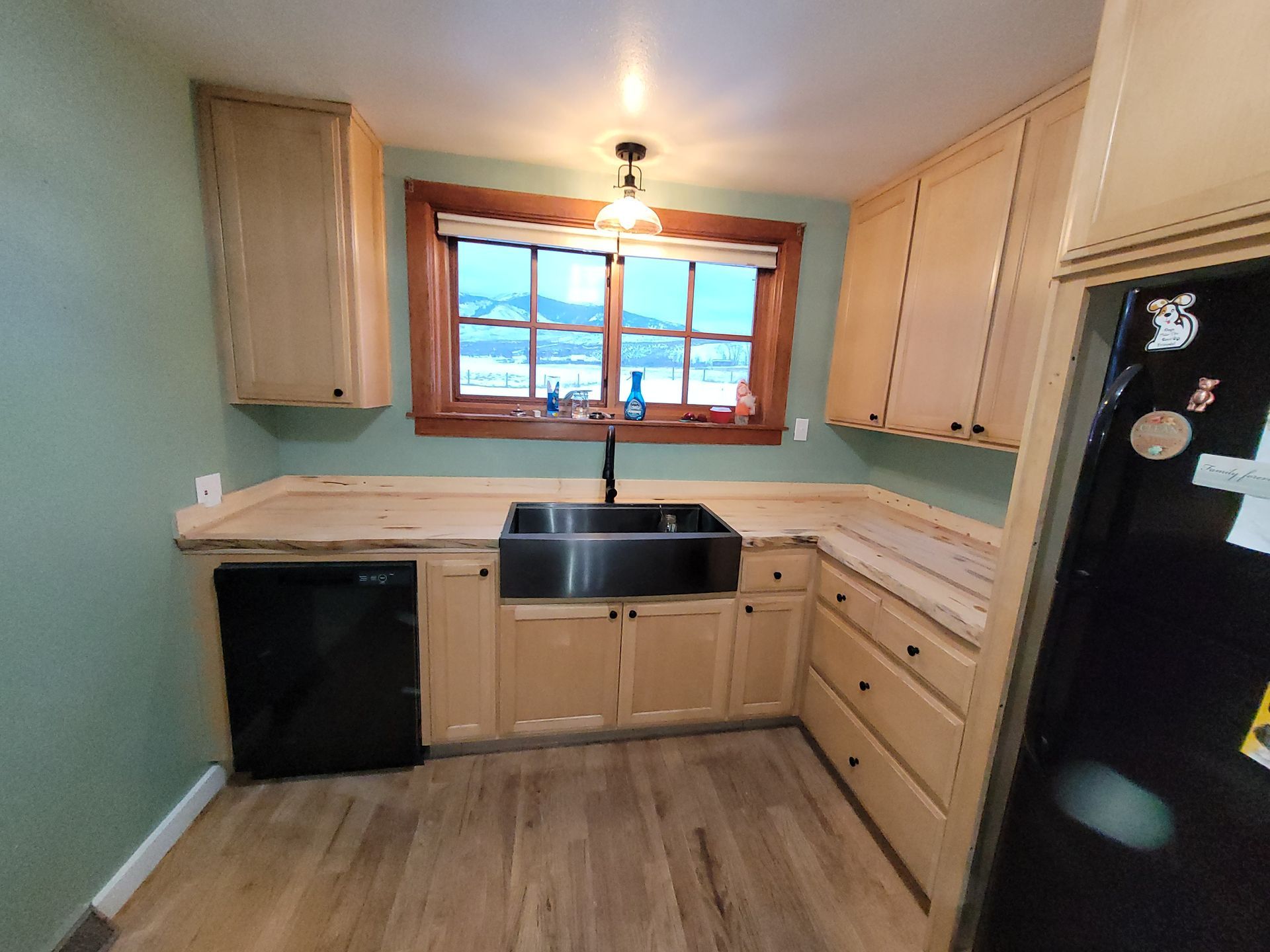A kitchen with light wood cabinets, light countertops, a black stainless steel sink, and a black dishwasher. A refrigerator is on the right, and a window with a mountain view is above the sink.