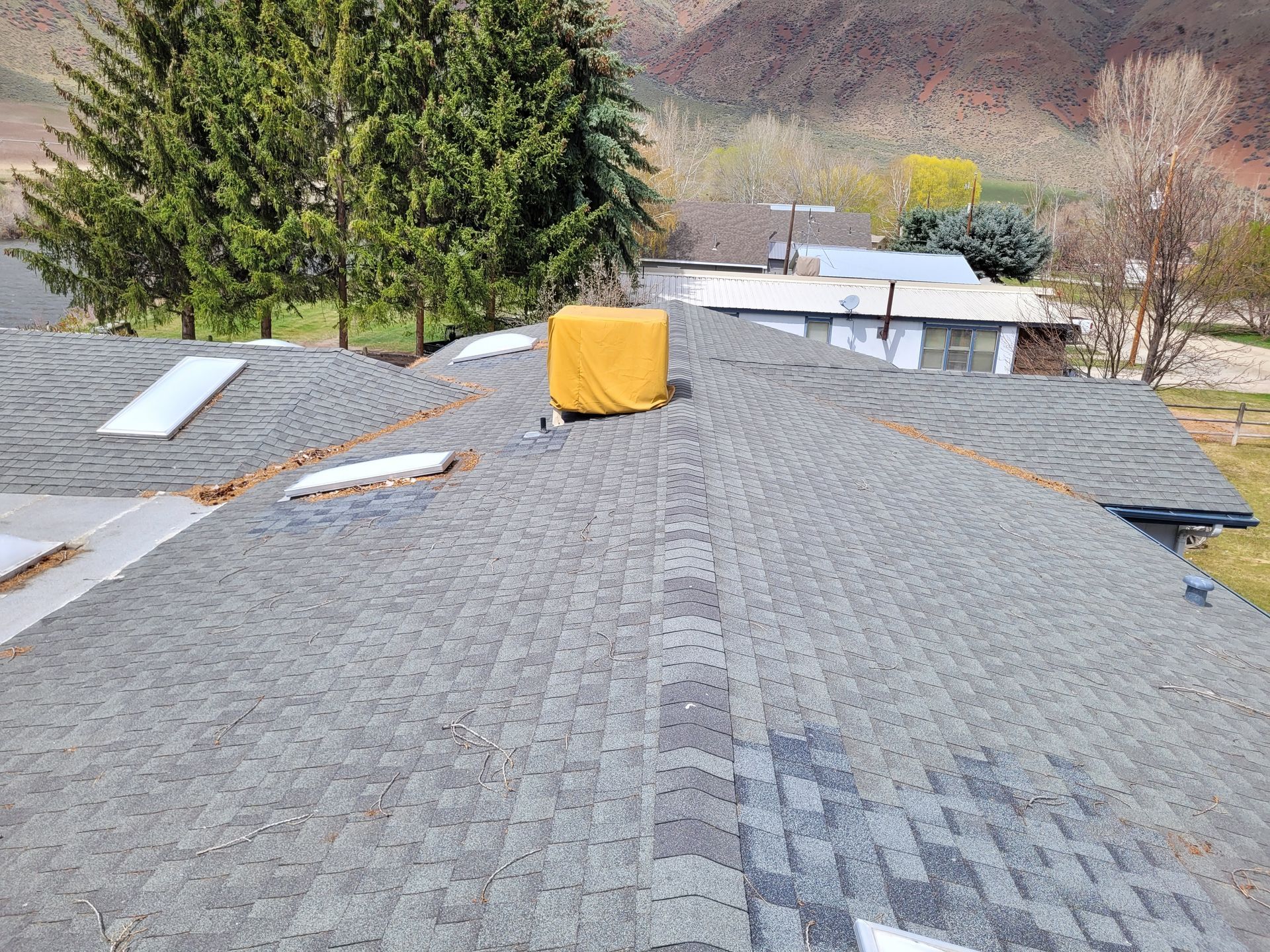 Rooftop with gray shingles, a yellow cover over a vent, and some missing shingles. Trees and a distant house are in the background.