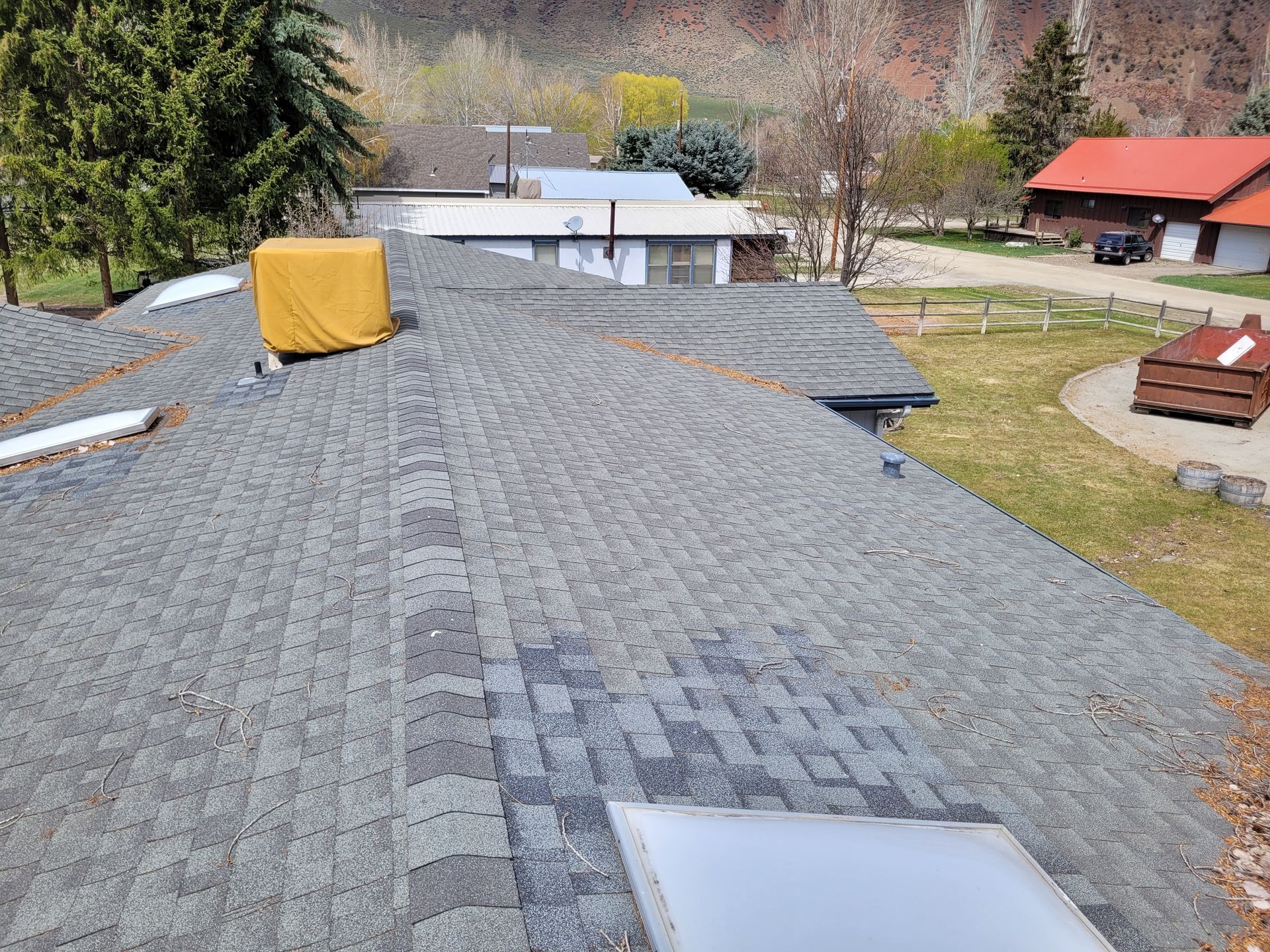 Gray shingled roof with a covered object, set against a mountain and building backdrop.