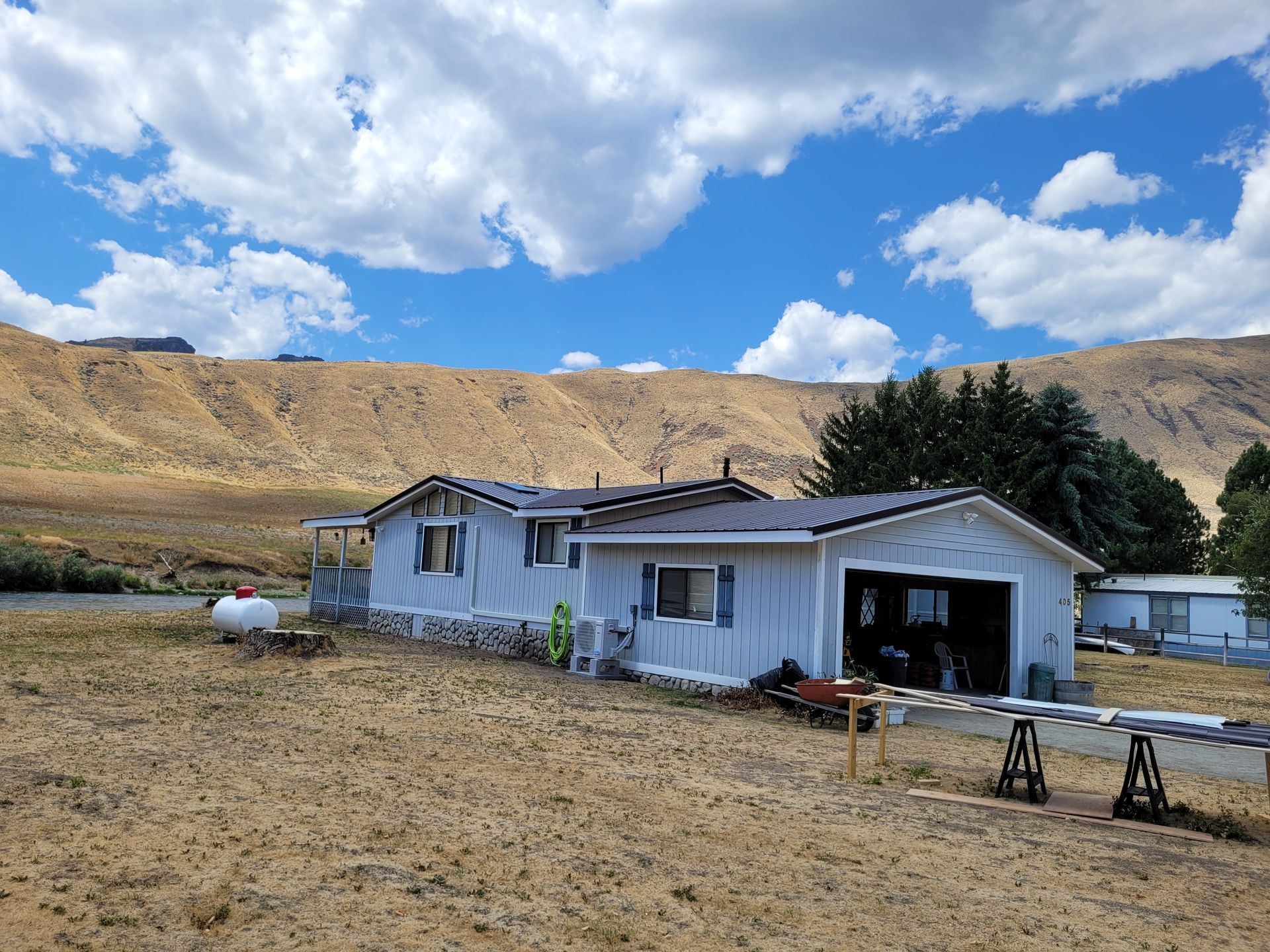 A light blue house with a garage, set in a dry field with a mountain backdrop under a cloudy sky.