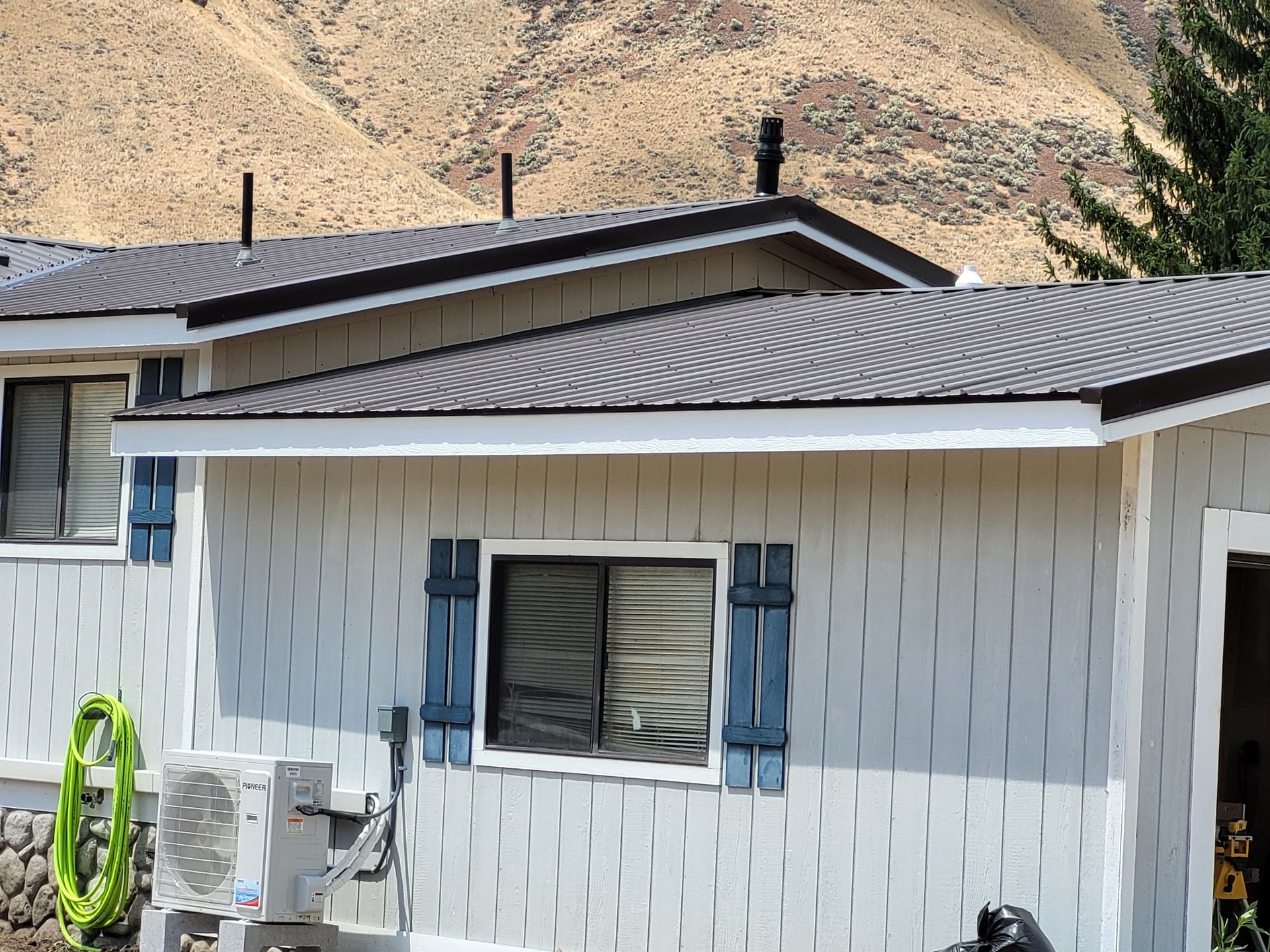 Light blue house with a dark roof, blue shutters, and a mountain backdrop. An air conditioning unit is visible.