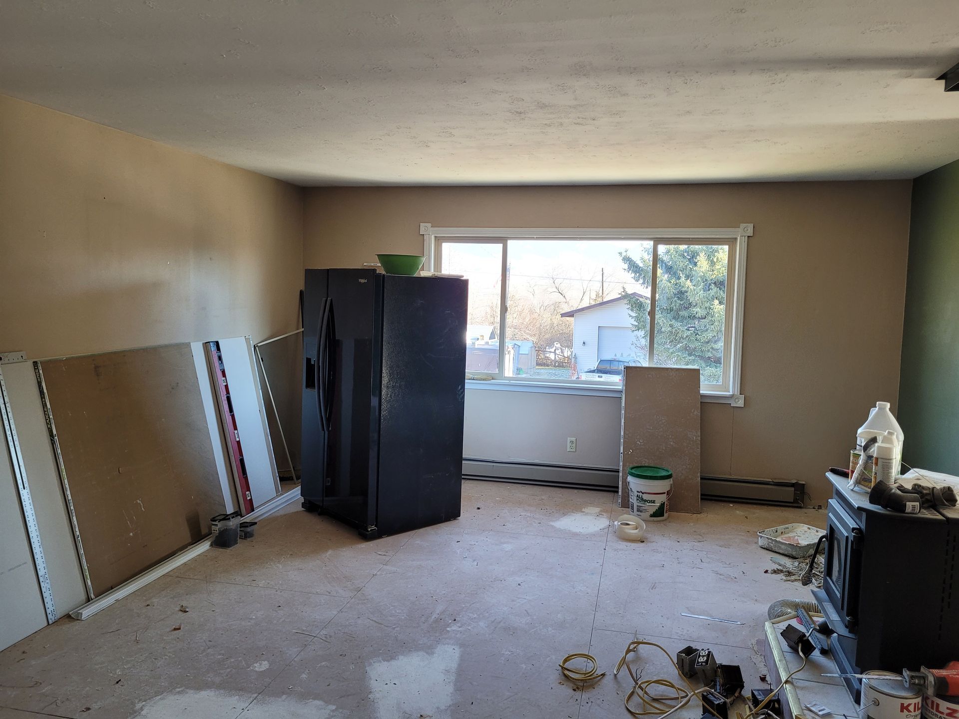 A room under renovation with drywall, an appliance, and tools scattered on the floor. Natural light streams through a window.