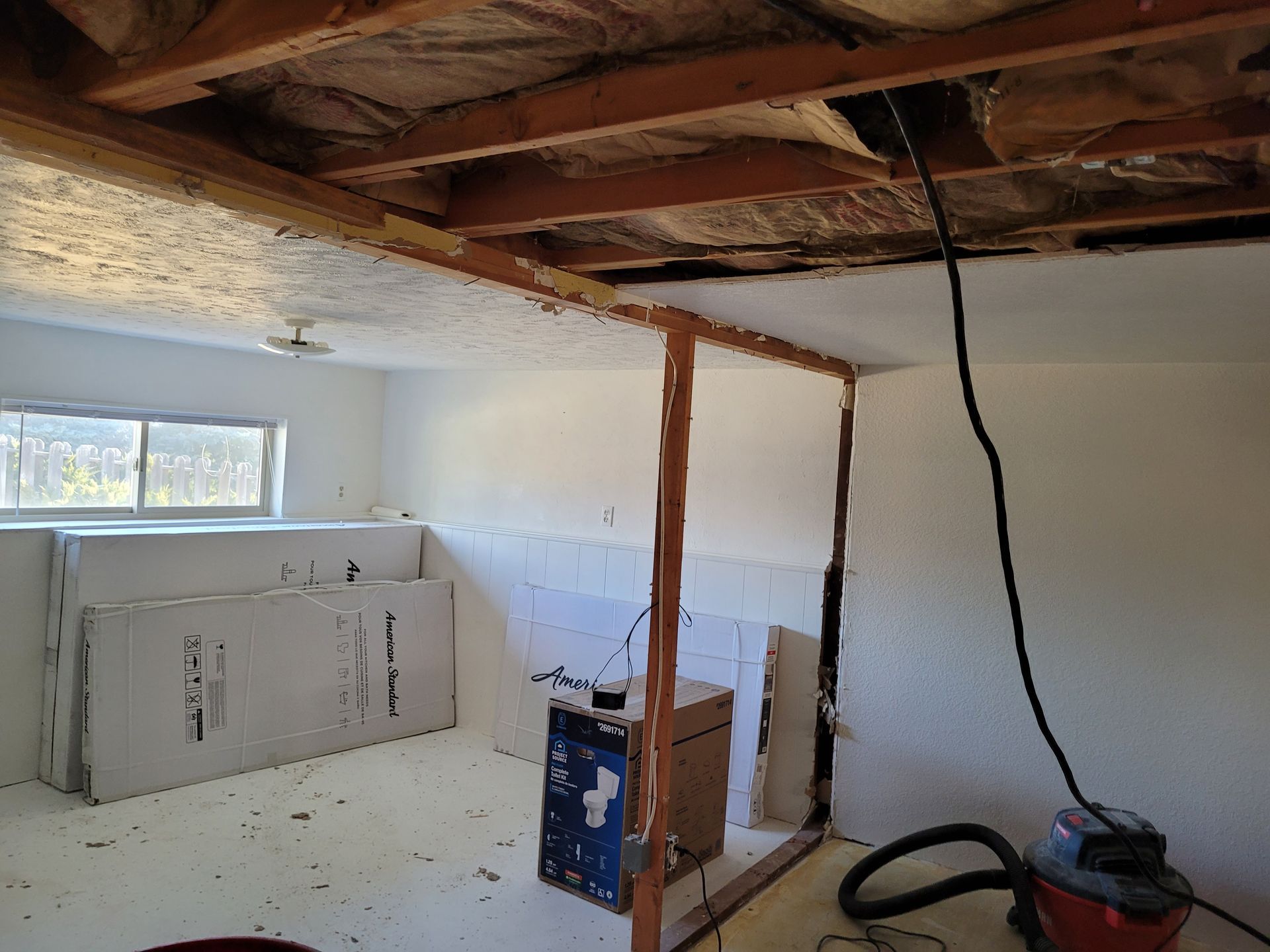 Interior view of a room under renovation with exposed wooden beams, a partial wall frame, and construction materials. A vacuum cleaner sits on the floor.