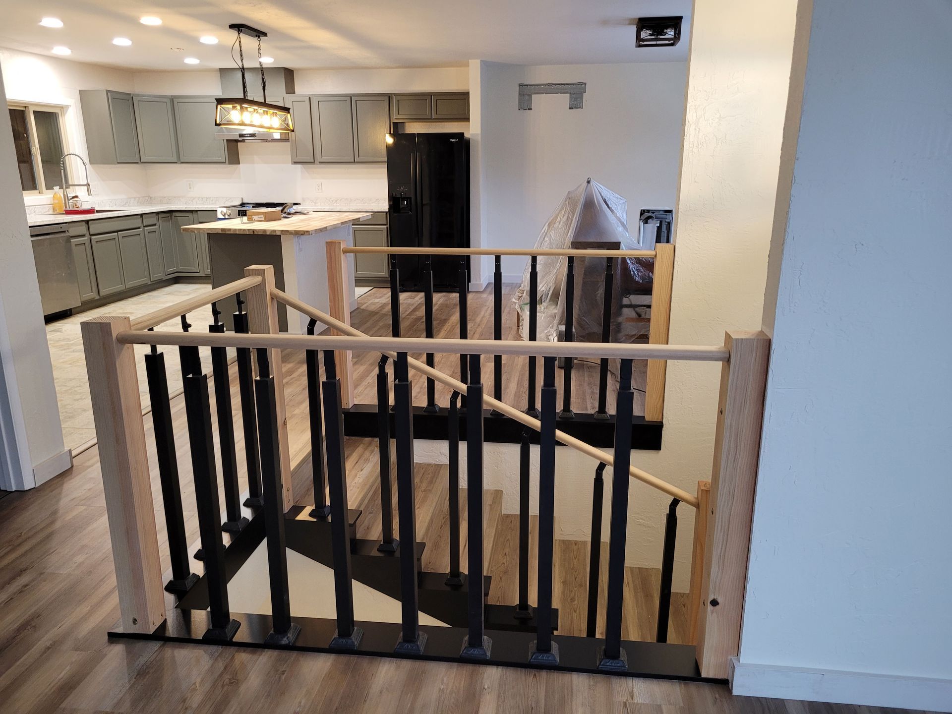 Interior view of a newly renovated home with a staircase featuring black vertical spindles and light-colored wooden handrails leading down to the kitchen area.
