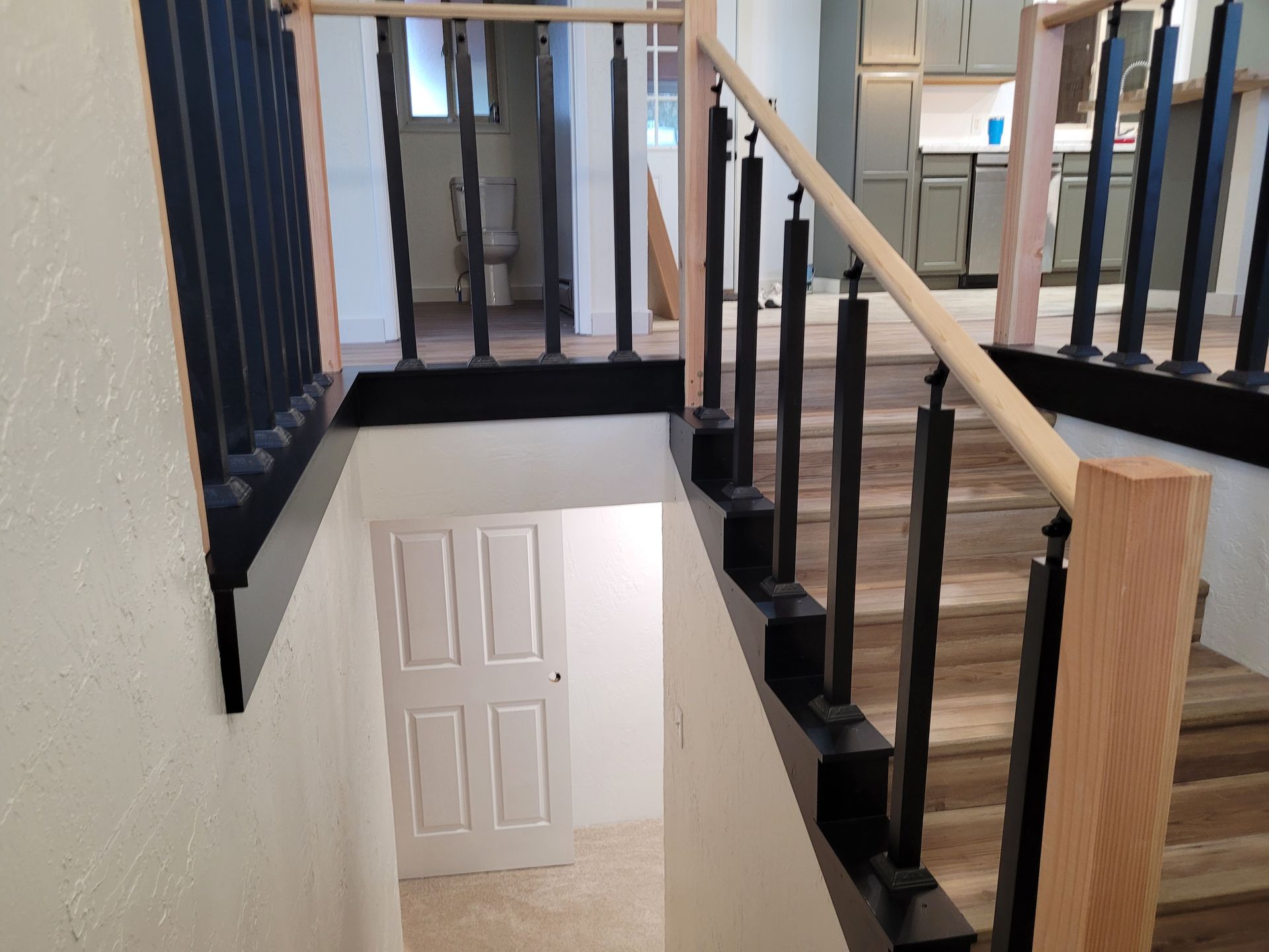 Staircase with black balusters, light wood railing, and a white door below.