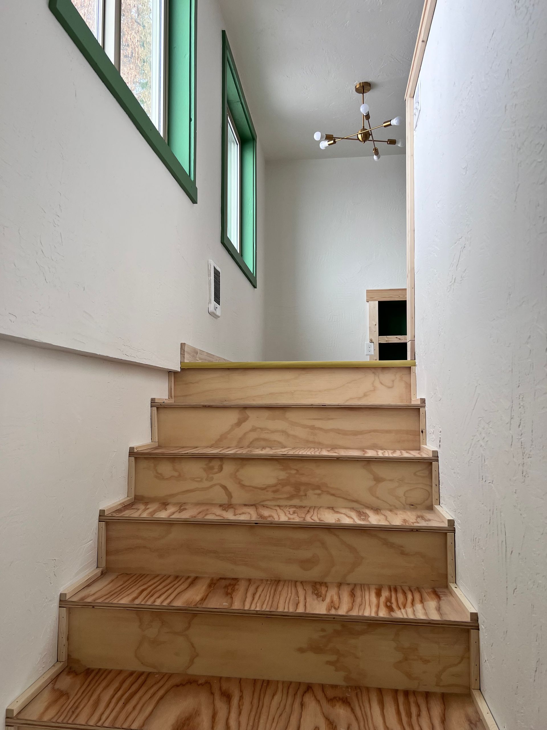 Staircase with plywood steps leading upwards, flanked by white walls and green window frames. A small chandelier hangs from the ceiling.