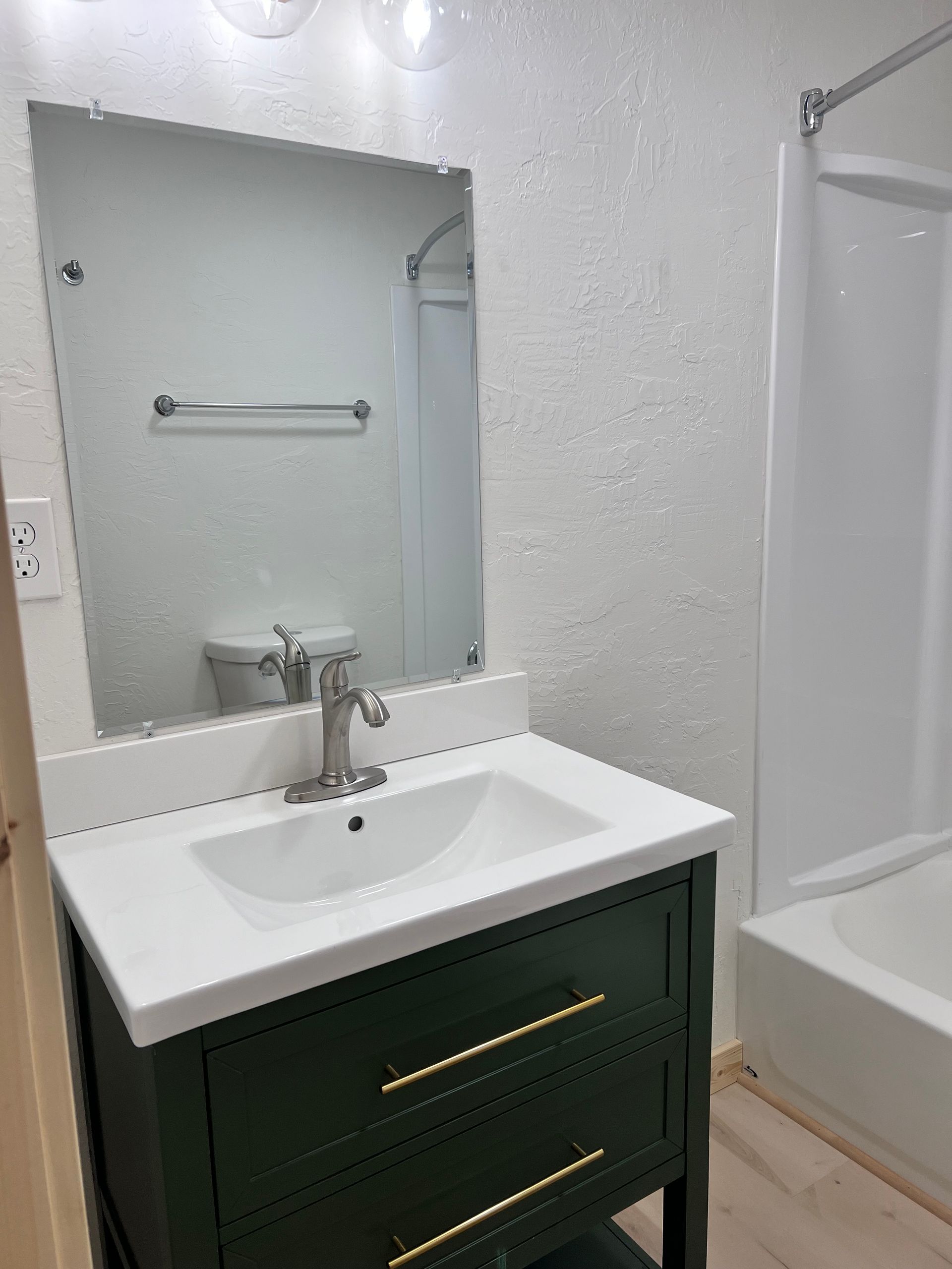 A renovated bathroom with a green vanity, white countertop, and a large mirror reflecting the toilet and shower.