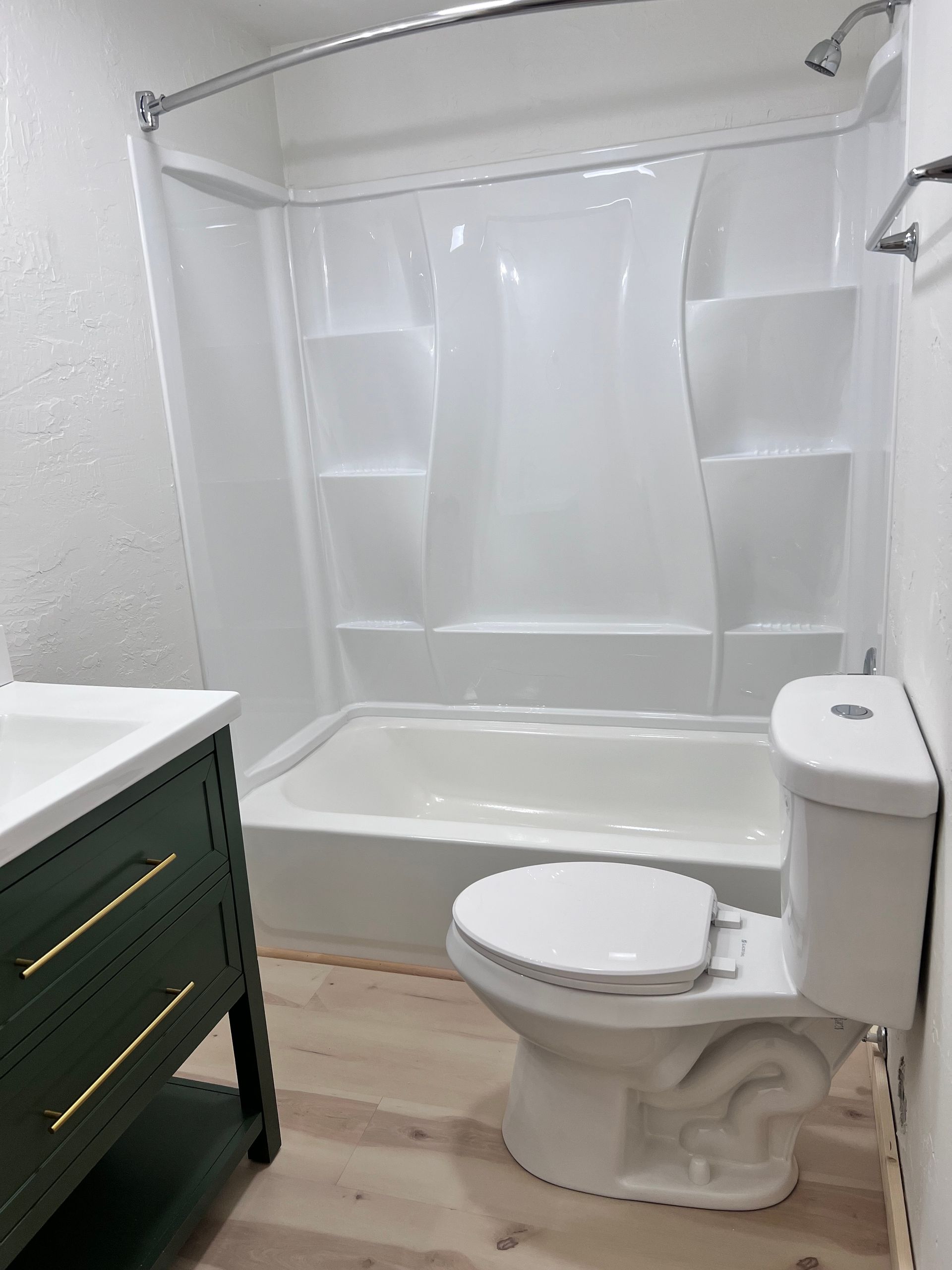 Small bathroom with a white tub/shower, toilet, and green vanity. Light wood-look flooring.