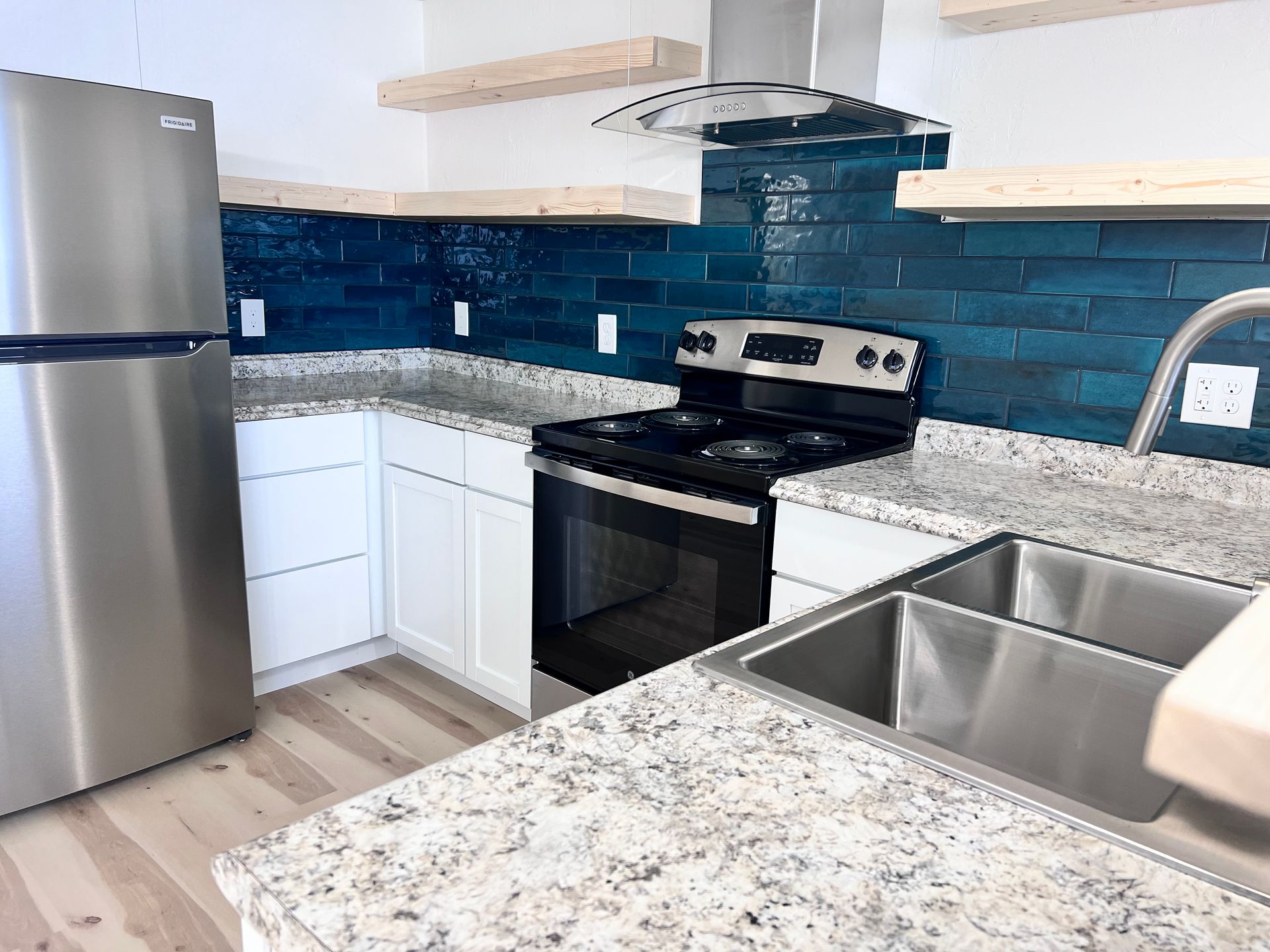 Modern kitchen with stainless steel appliances, white cabinets, and a blue tile backsplash. It features light-colored countertops and wooden shelving.