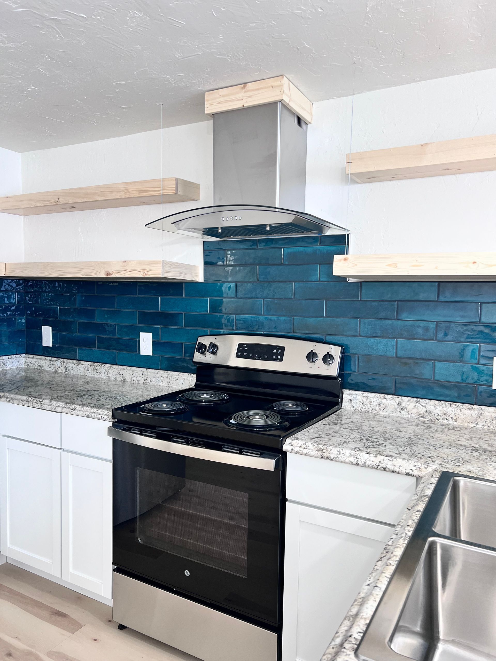 Modern kitchen with white cabinets, granite countertops, teal backsplash, stainless steel range, and vent hood. Wooden shelves are mounted on the white walls.