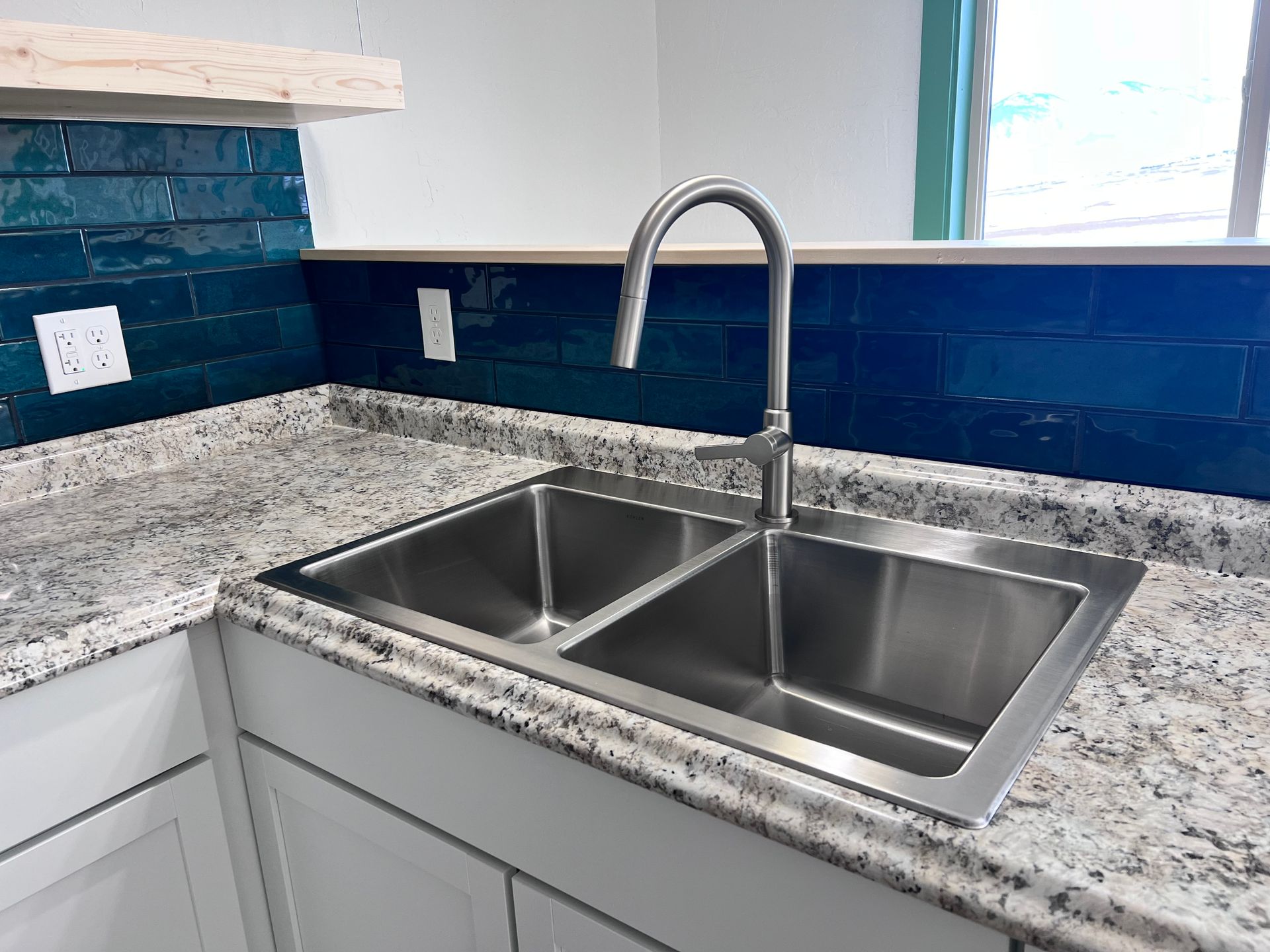 Stainless steel double sink in a kitchen with granite countertops and blue tiled backsplash.