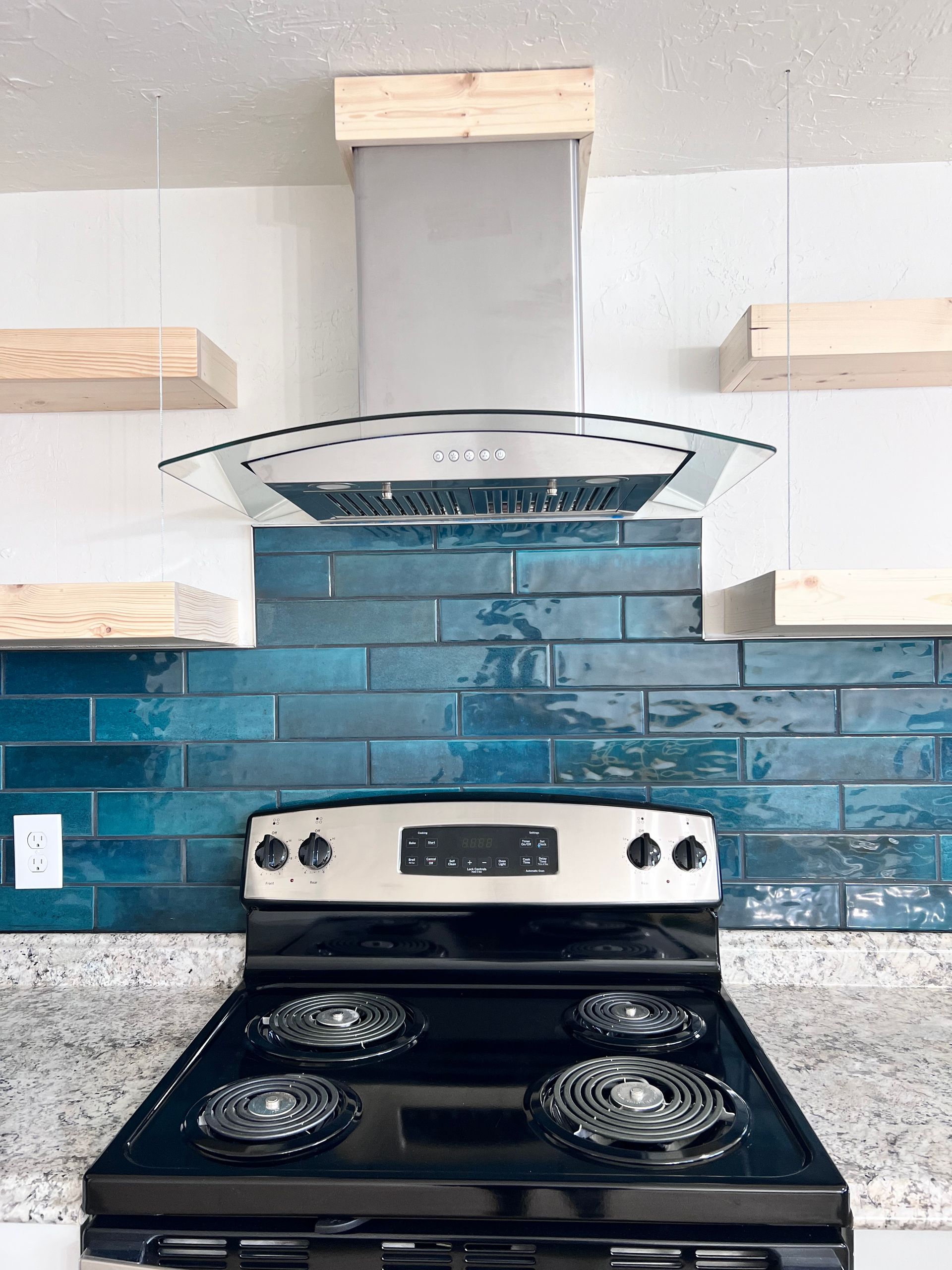 Kitchen with a stainless steel range hood over a black electric stove and teal blue tiled backsplash. Floating wooden shelves are on either side.
