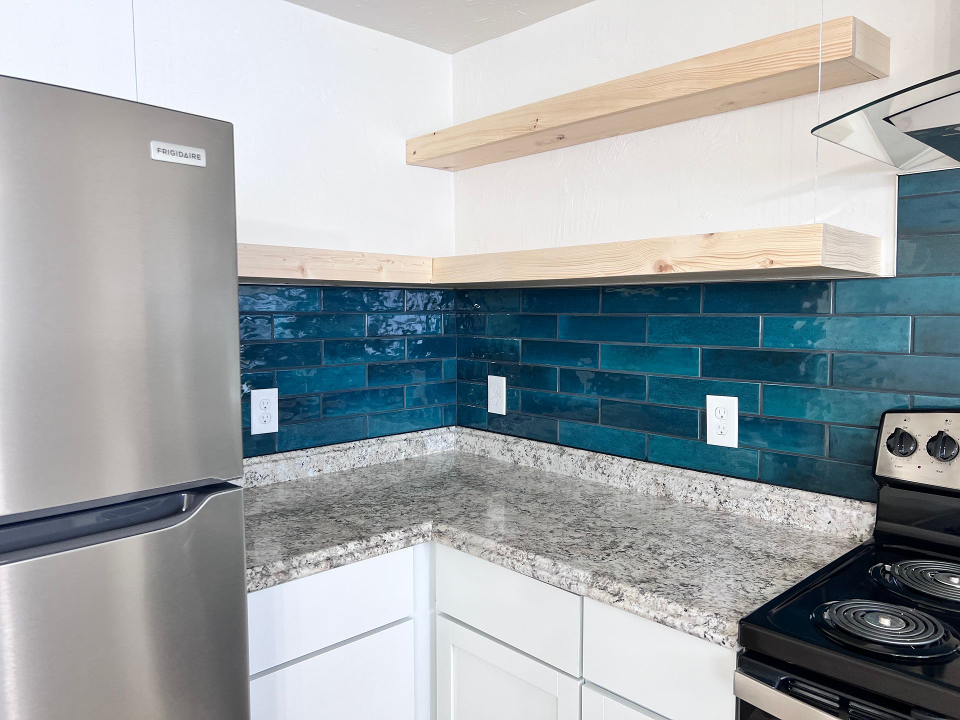 Kitchen corner with teal tiled backsplash, granite countertop, and wooden floating shelves. Stainless steel fridge and range.