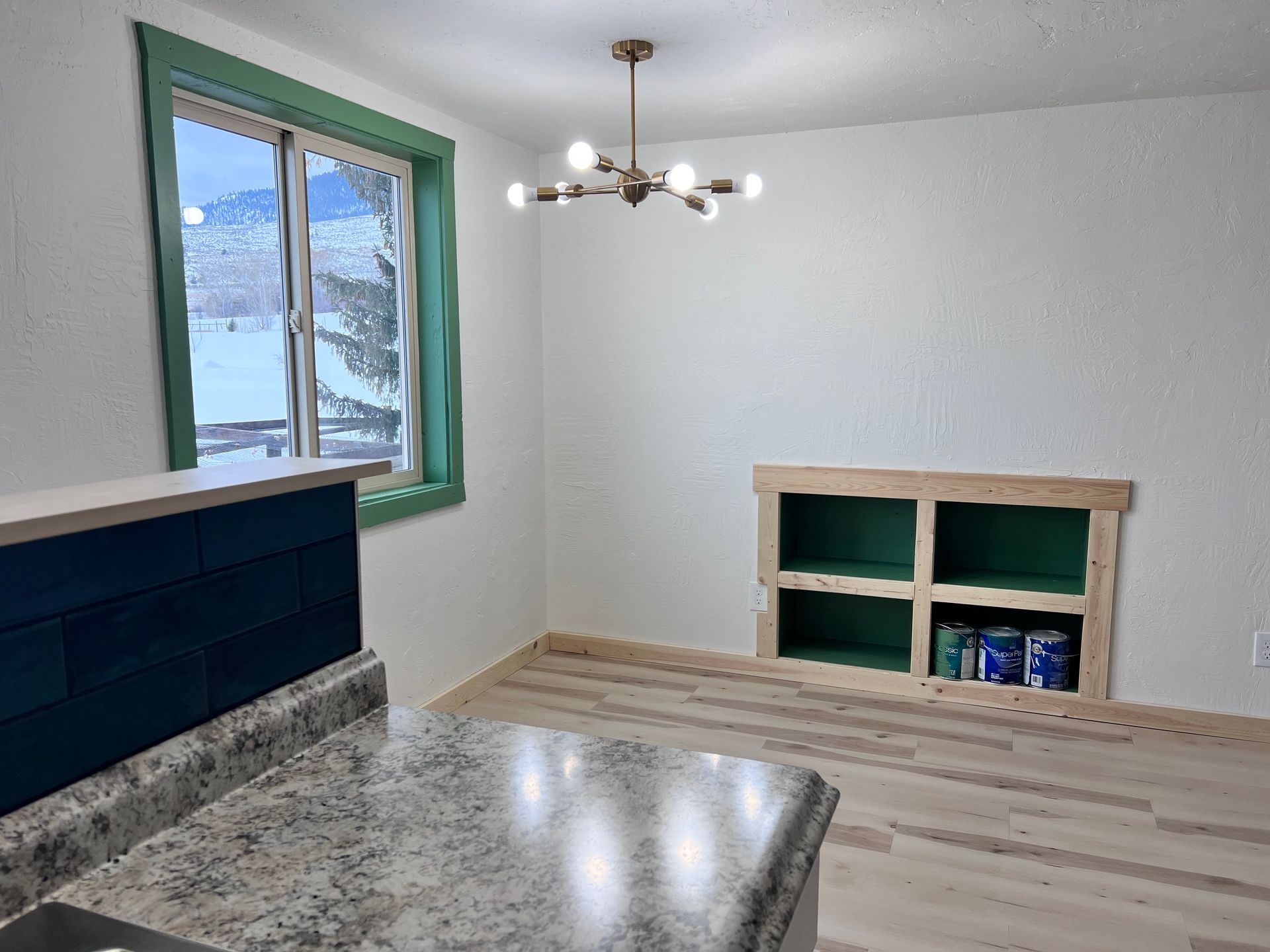 A dining area with white walls, a green-framed window, and a Sputnik-style light fixture. A built-in shelf is painted green.