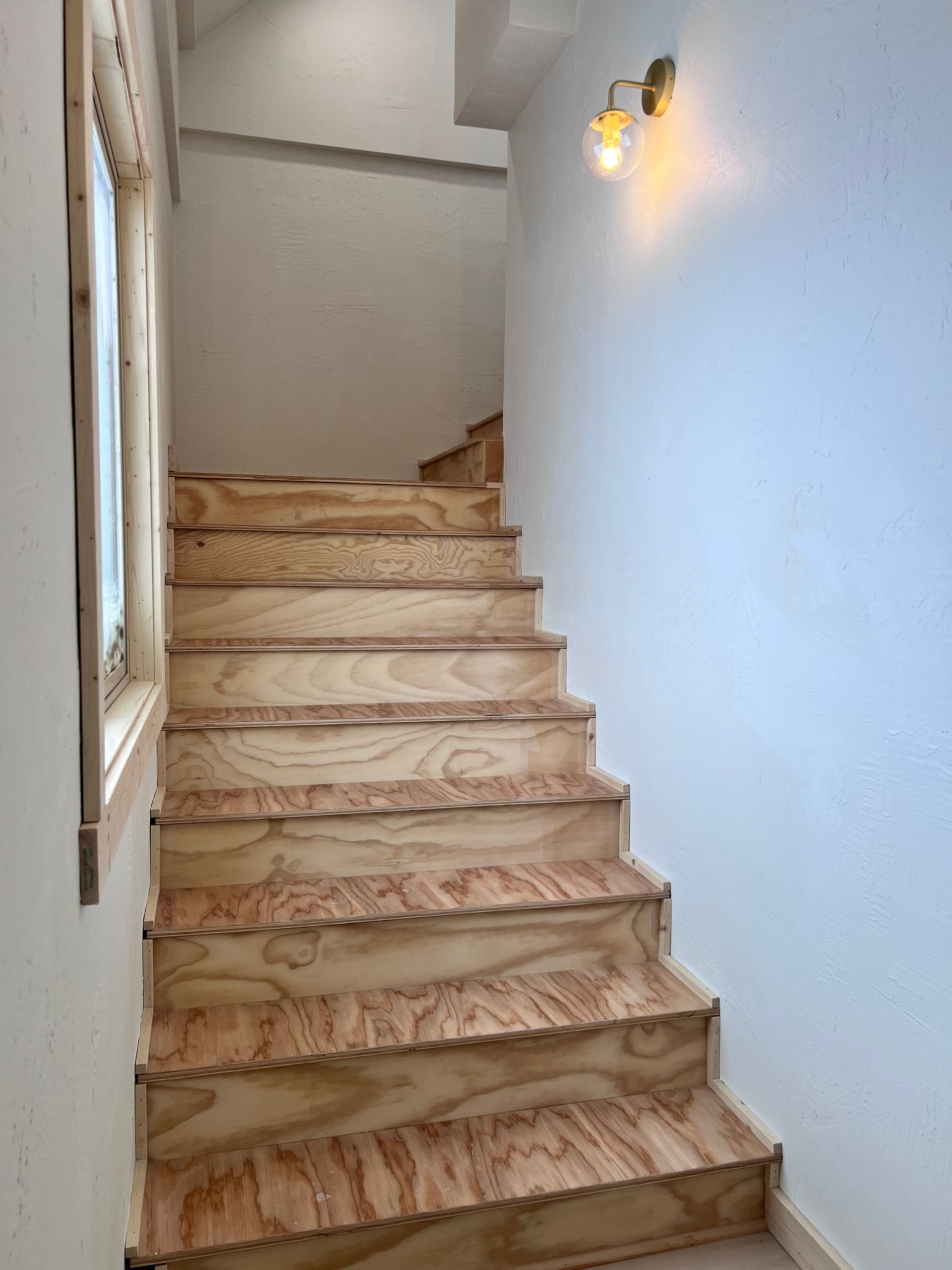 Staircase with light wood steps and a wall-mounted light fixture. There is a window to the left and a blue wall.