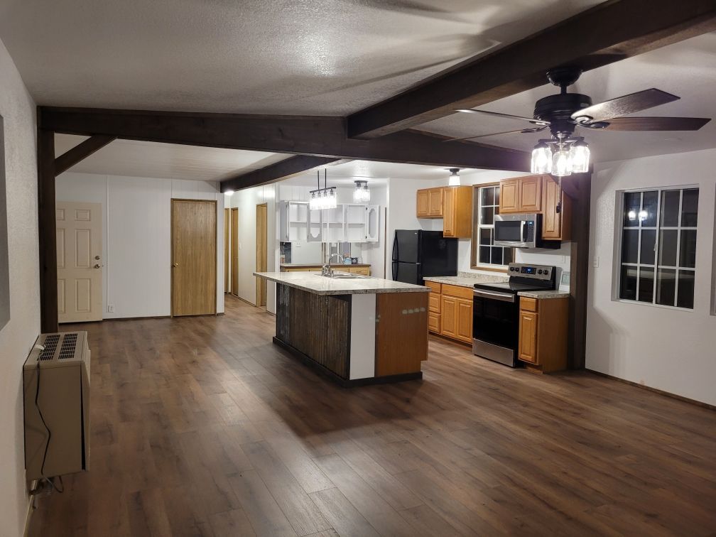 Open-concept kitchen with a large island, dark appliances, wood cabinets, and sliding glass door. Wood floors and ceiling beams complete the space.
