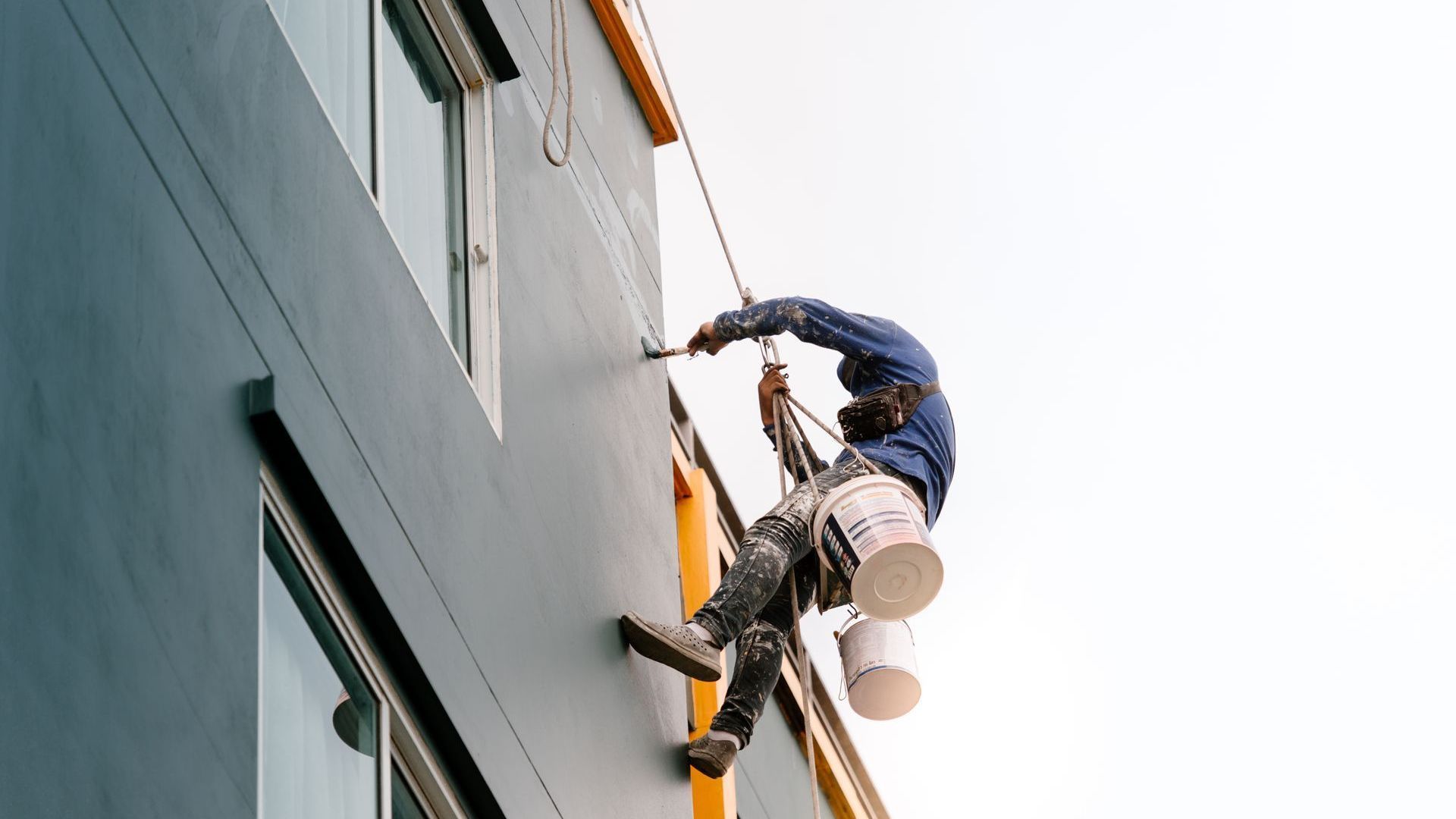 A man is painting the side of a building on a rope.