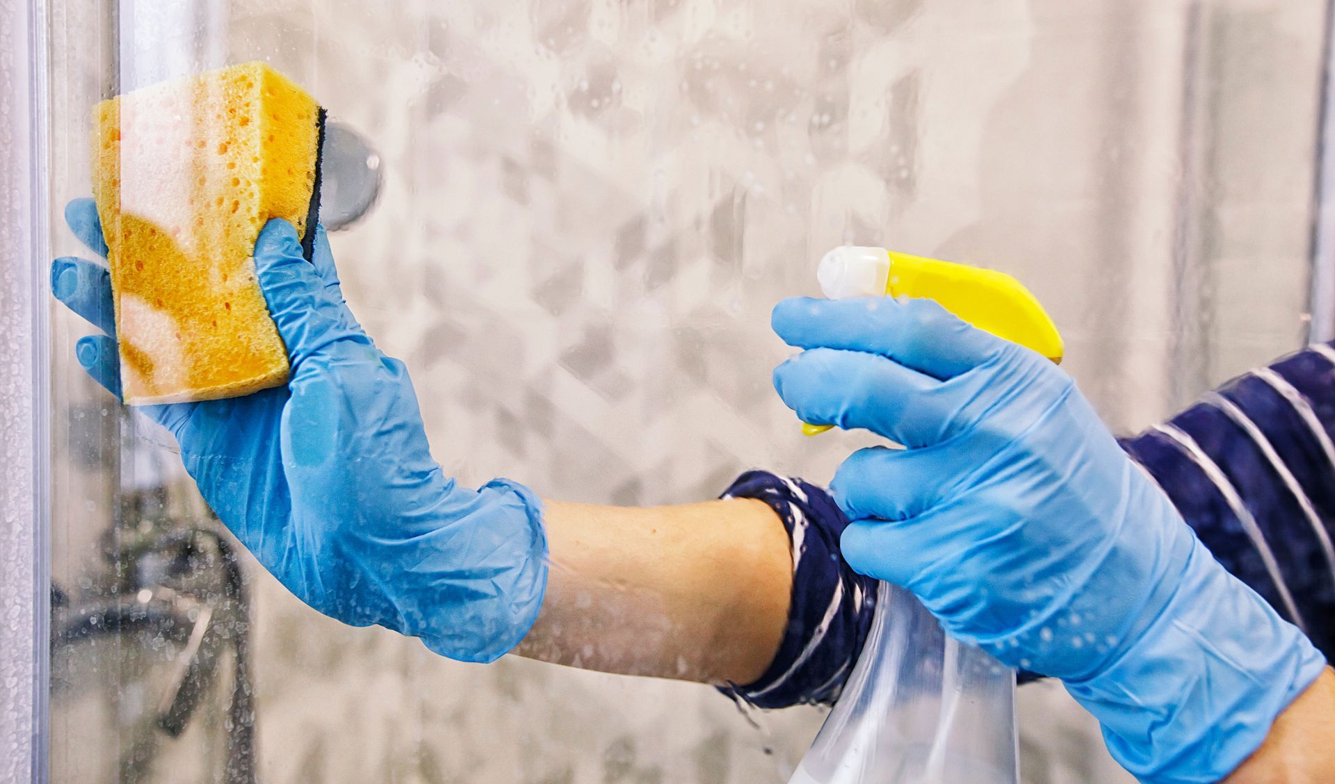 A person wearing blue gloves is cleaning a shower door with a sponge and spray.