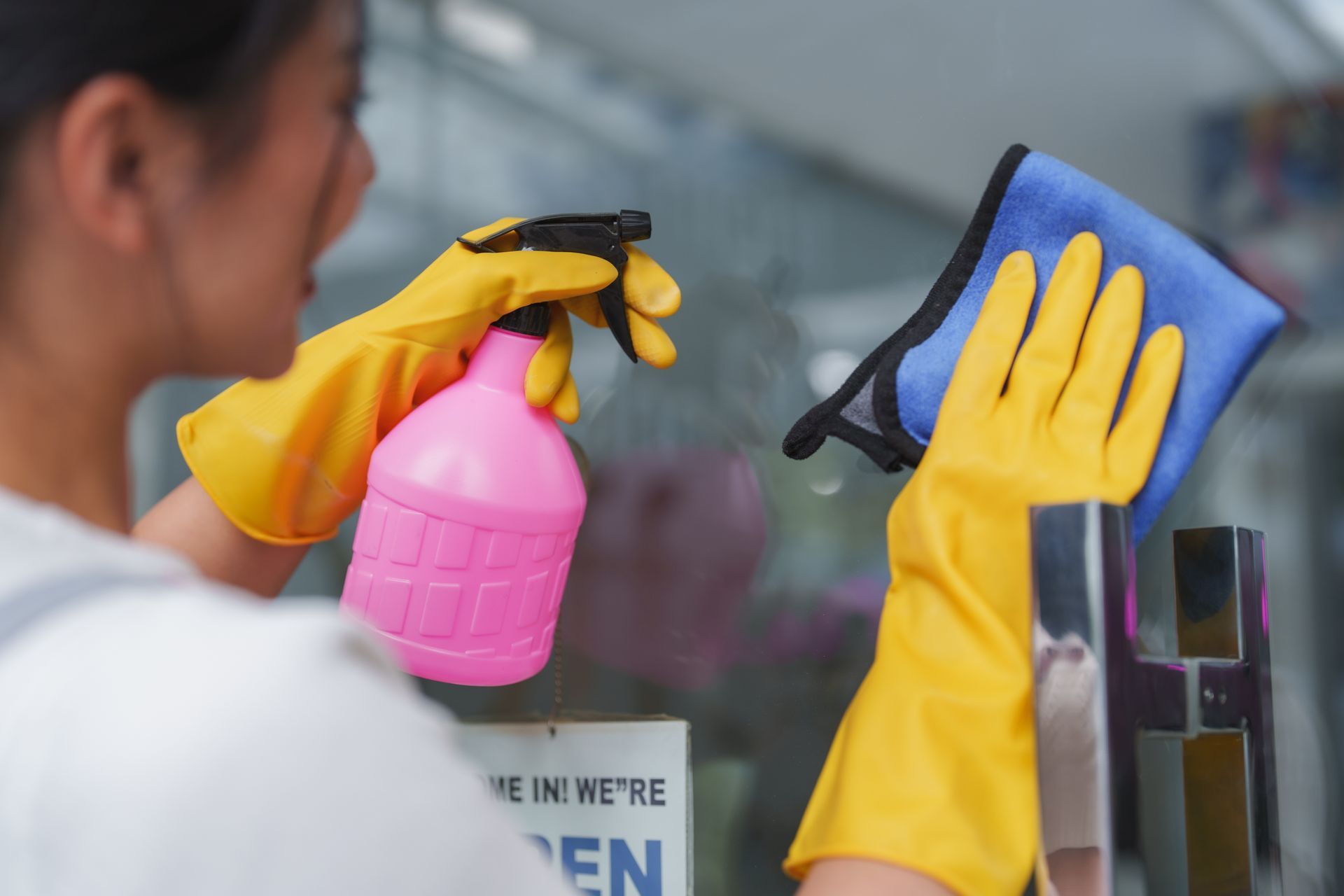 A woman wearing yellow gloves is cleaning a window with a spray bottle and a cloth.