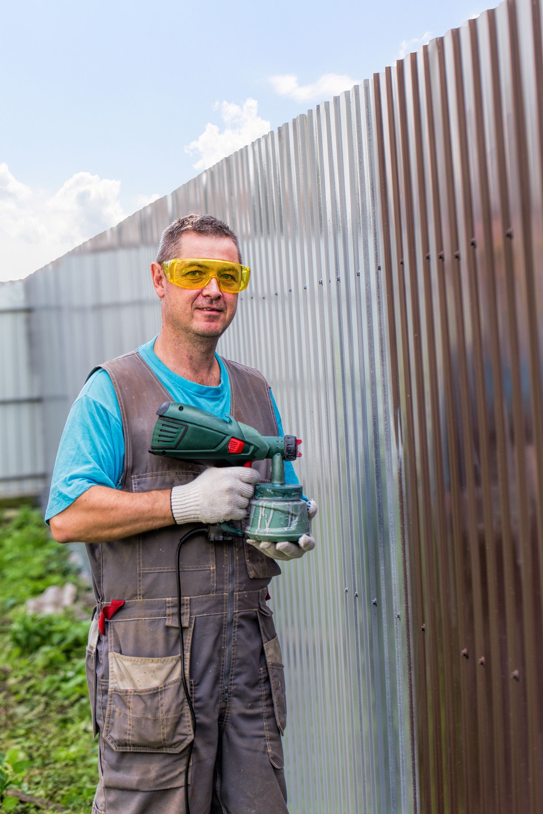 A man in overalls and protective glasses with spray gun in hand applying water resistant coating on a metal fence