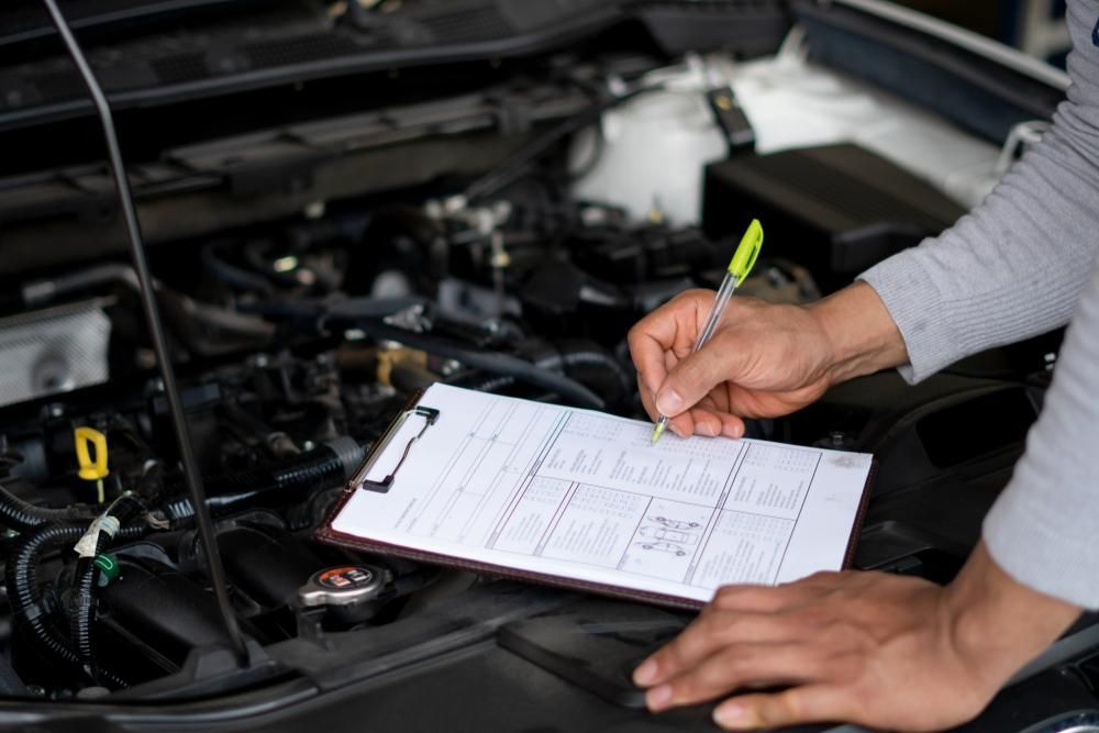 A Man Is Writing on A Clipboard in Front of A Car Engine — Mike's Automotive in Edgeworth, NSW