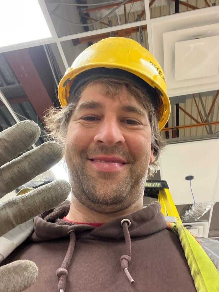 Man in yellow hard hat, brown hoodie, and work gloves smiles, waving. Construction setting.
