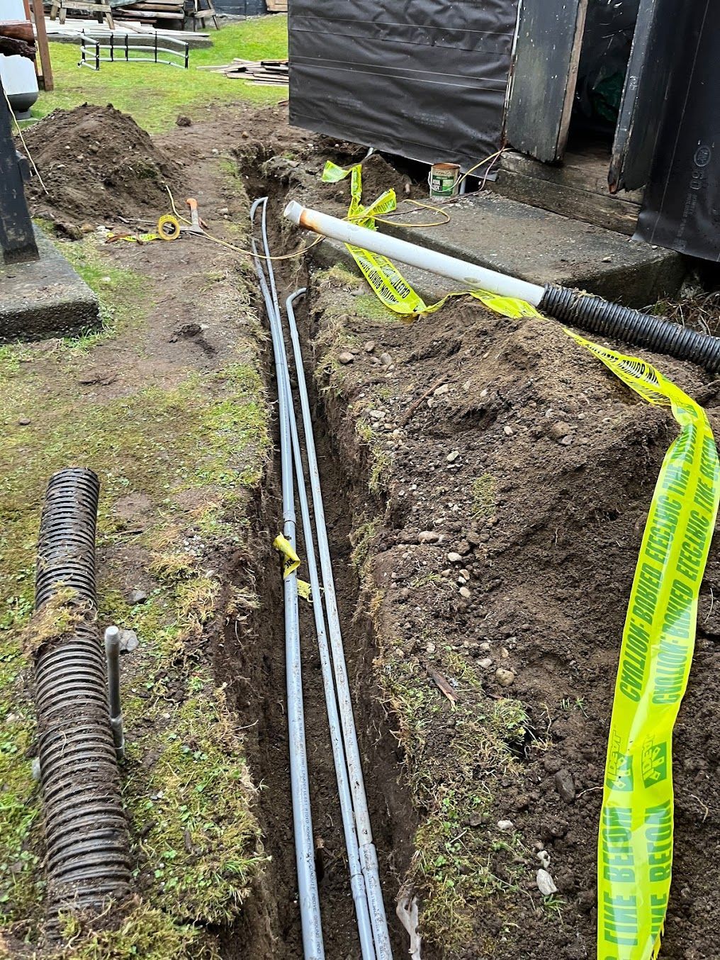 Trench with gray pipes, yellow caution tape, and dirt, likely for underground utilities.