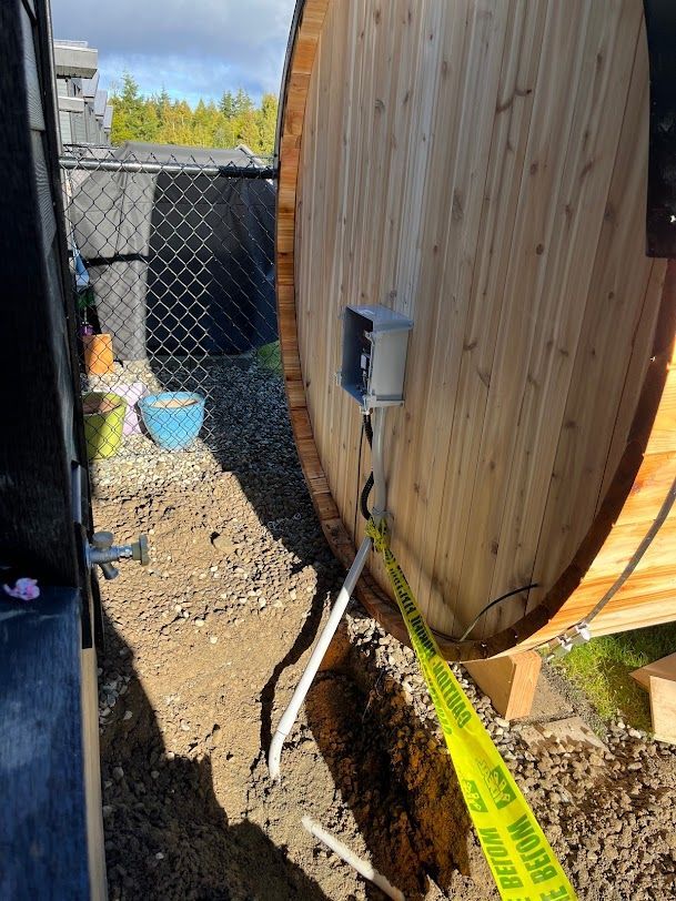 Wooden sauna barrel with electrical box, buried wire, and caution tape. Outdoors, next to a chain-link fence.