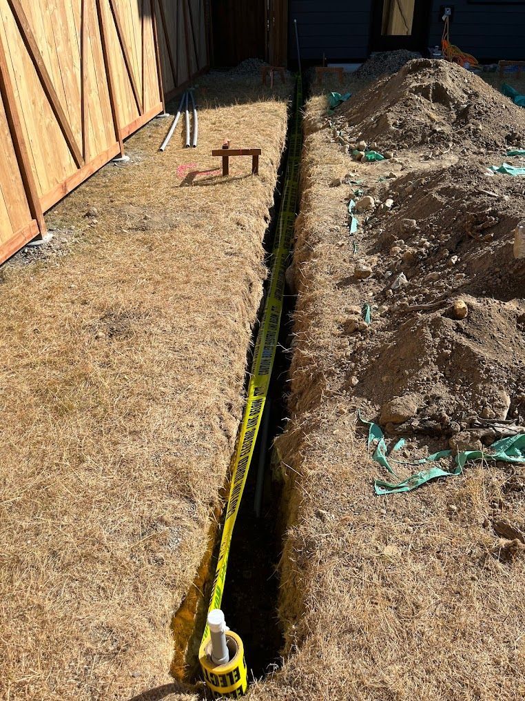 Narrow trench in yard with yellow caution tape, wood fence, and dirt piles.