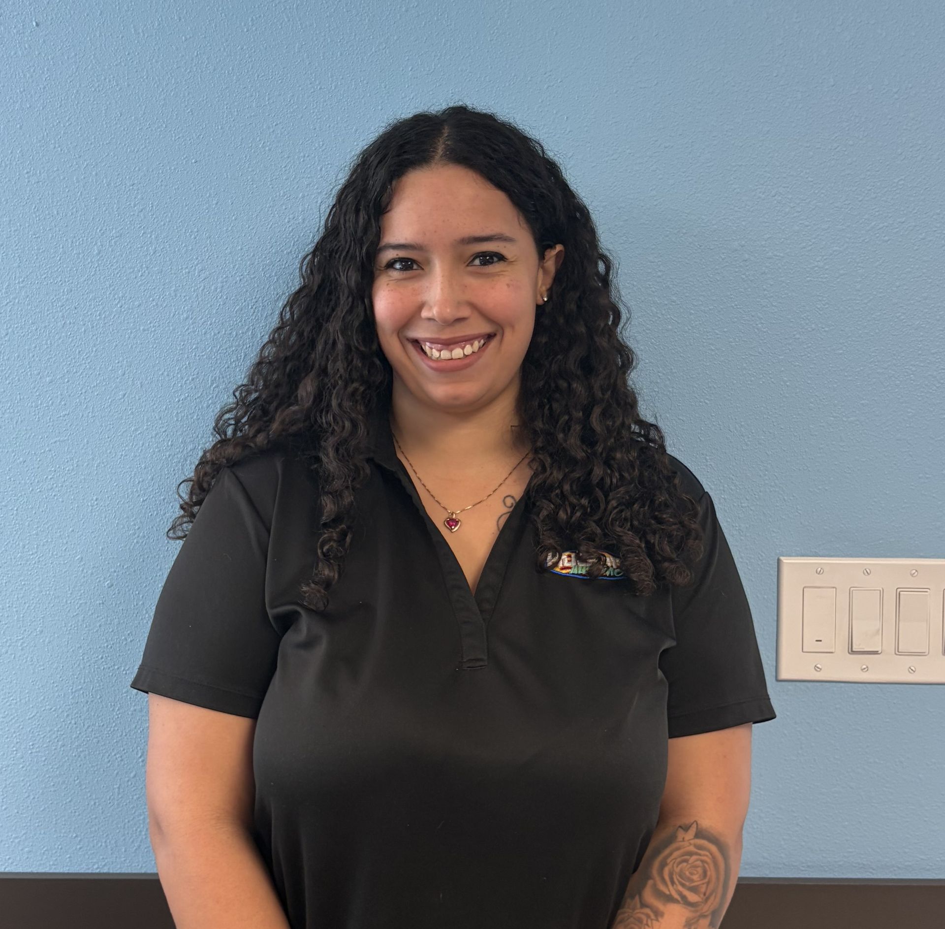 Woman with long curly hair smiles in front of a blue wall; wearing a black polo shirt and has a tattoo on her arm.