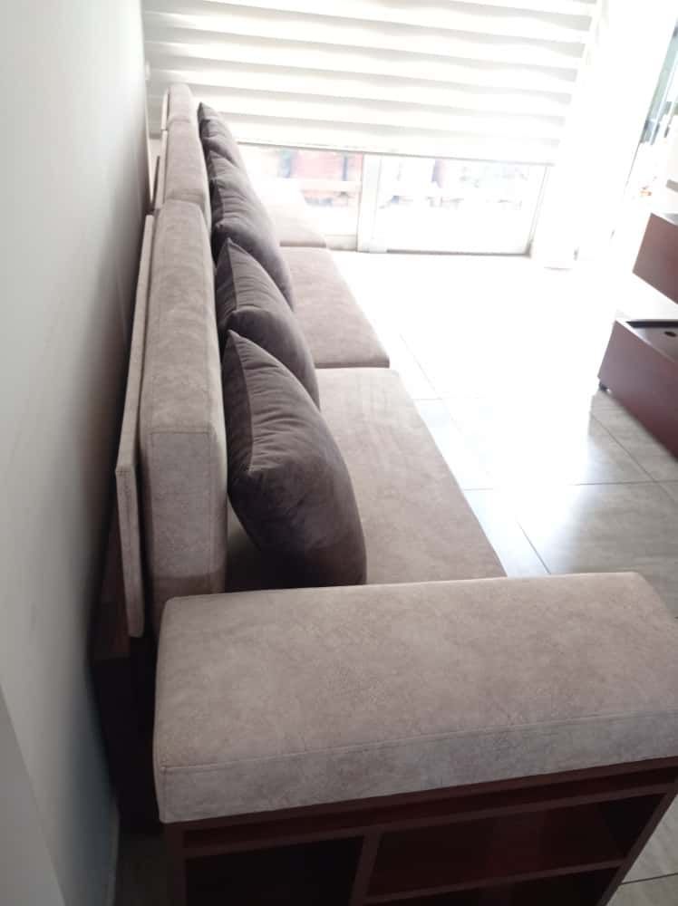 Sofá seccional beige con almohadas gris oscuro cerca de una ventana con persianas.