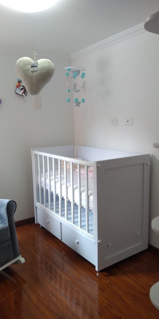 Una cuna blanca en la habitación de un bebé con suelo de madera, con un móvil en forma de corazón colgado encima.