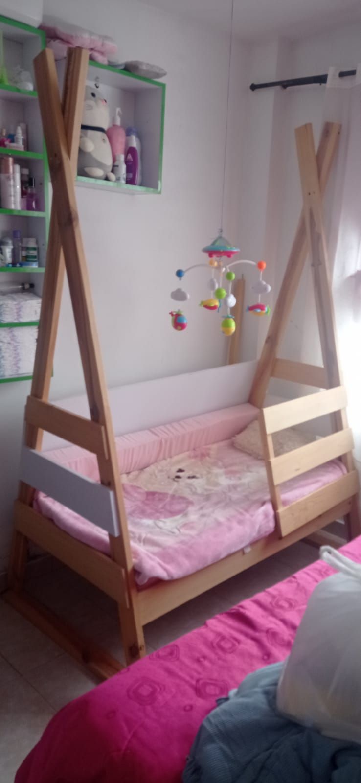 Cama estilo tipi de madera con ropa de cama rosa y móvil, colocada en una habitación con pared y estantería blancas.