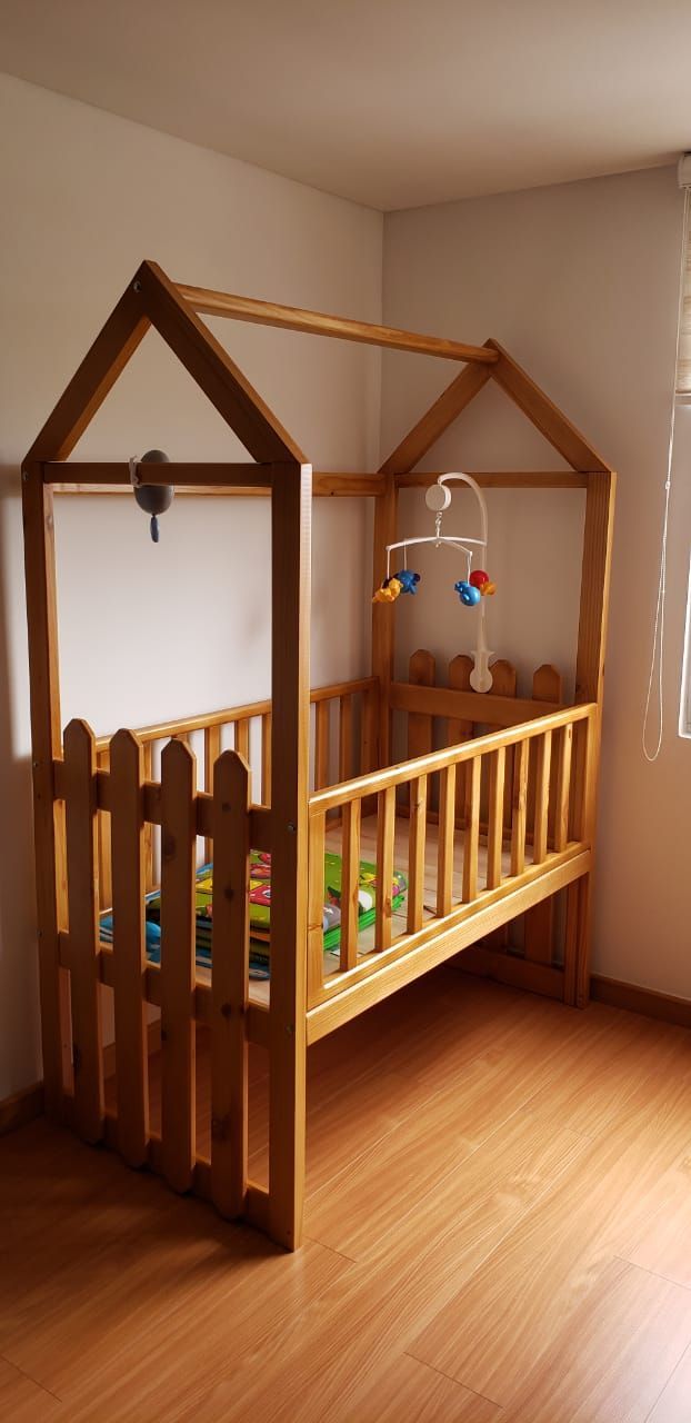 Cuna de madera con estructura en forma de casa y un móvil colgado en el interior. La cuna se asienta sobre un suelo de madera.