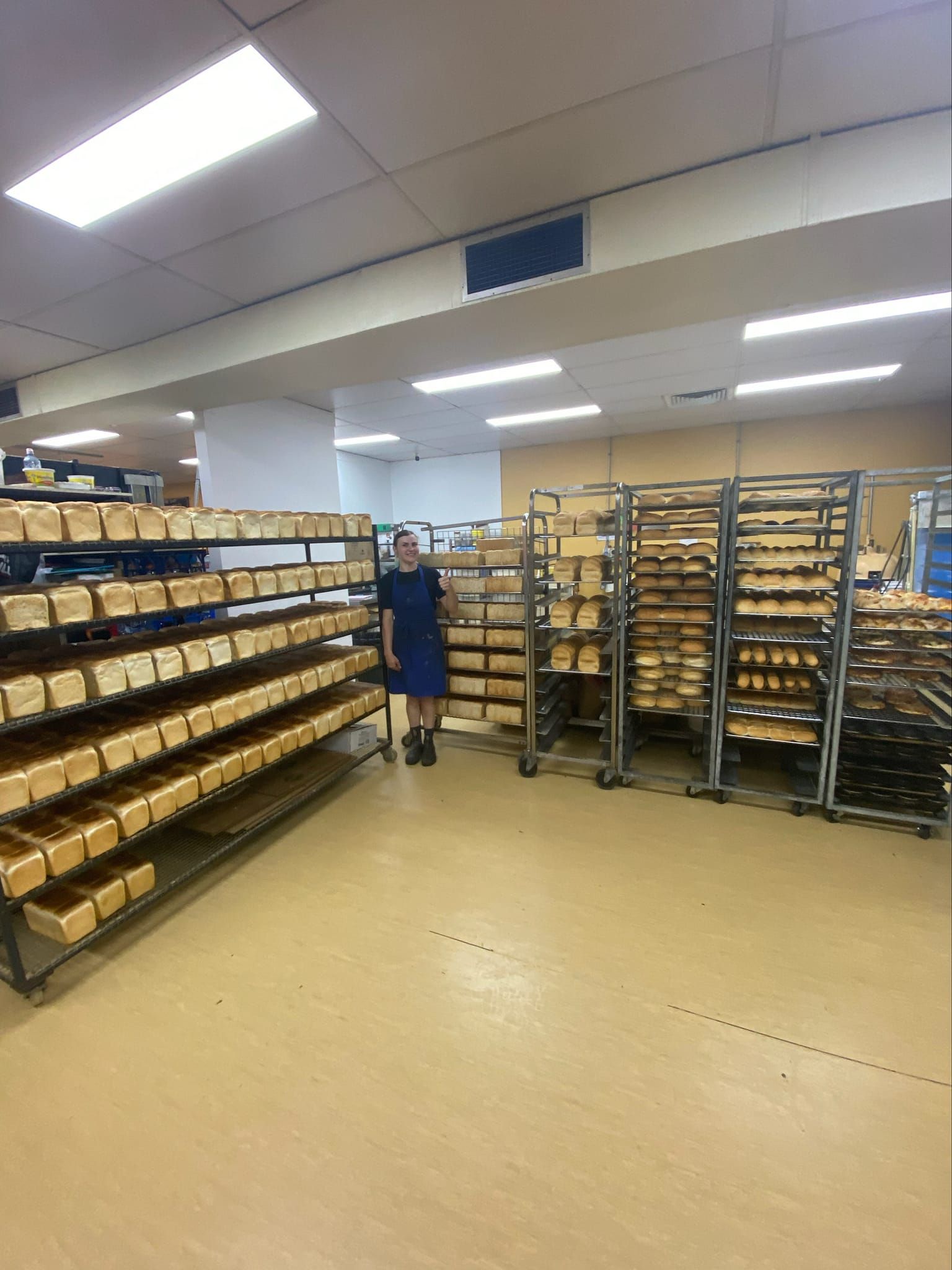 Person standing in a bakery with many shelves of bread. Bright lighting, tan floor — Atherton Bakehouse in Atherton, QLD 