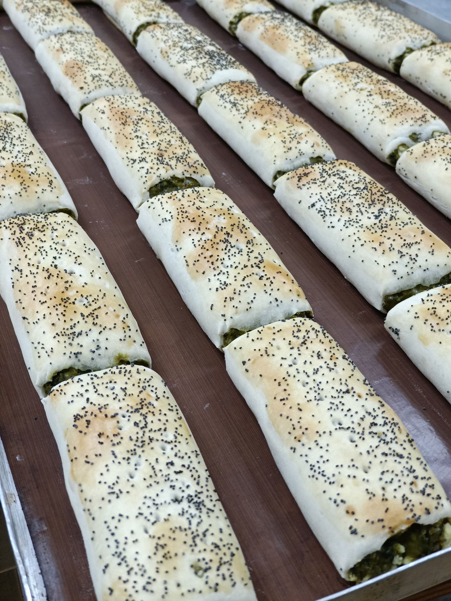Rows of baked pastries filled with green filling and topped with seeds on a baking sheet.— Atherton Bakehouse in Atherton, QLD