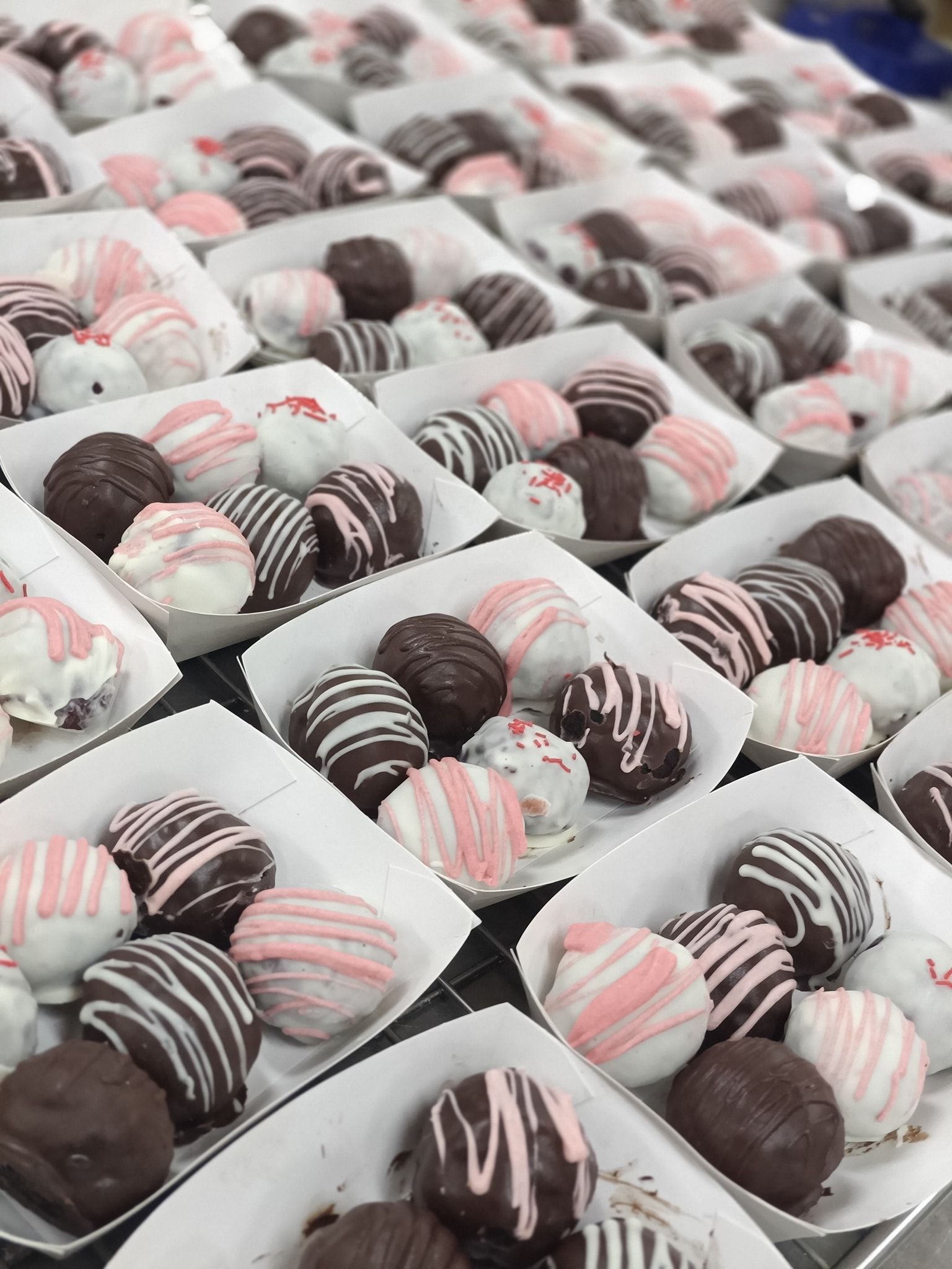 Rows of chocolate truffles in white paper trays, drizzled with white and pink icing 